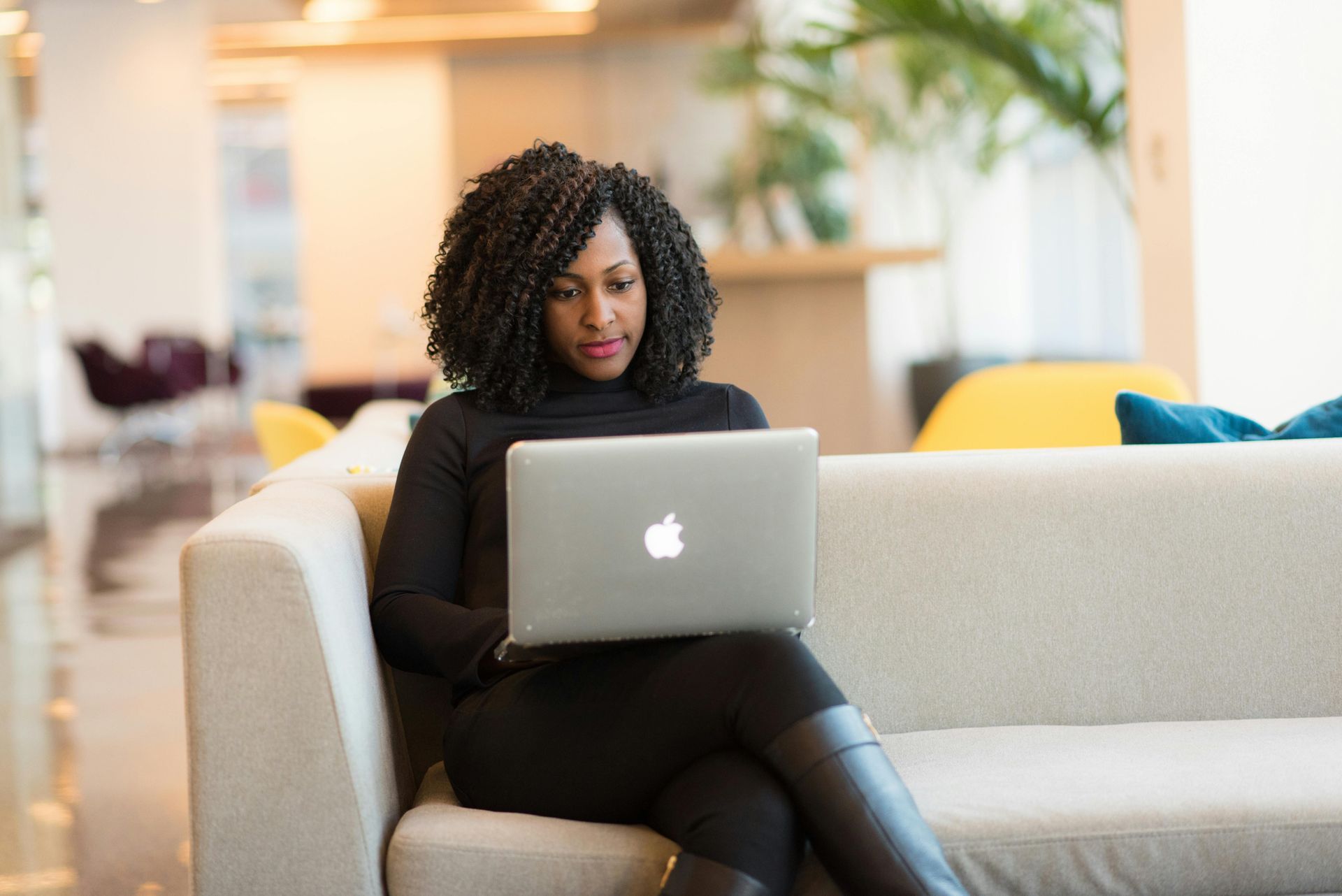 Woman using a laptop while sitting on a light-colored couch in a modern office.