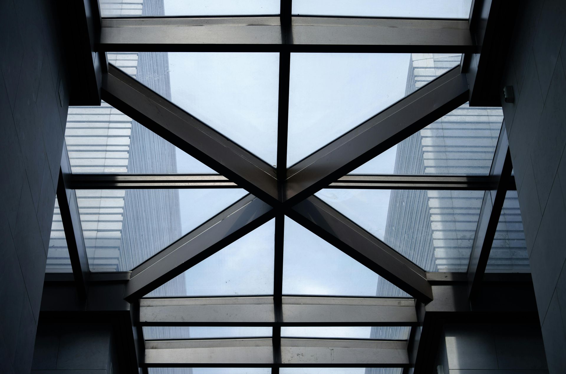 A glass ceiling with dark metal structural beams forming an X pattern, framed by tall, modern buildings.