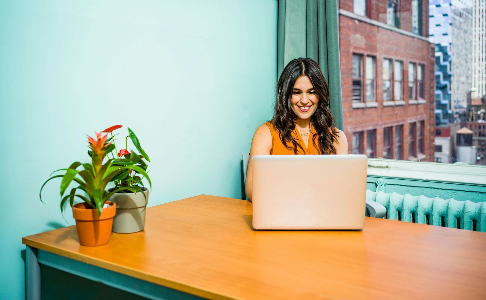 Woman smiling at laptop in an office setting, with a building view and plants on the desk.