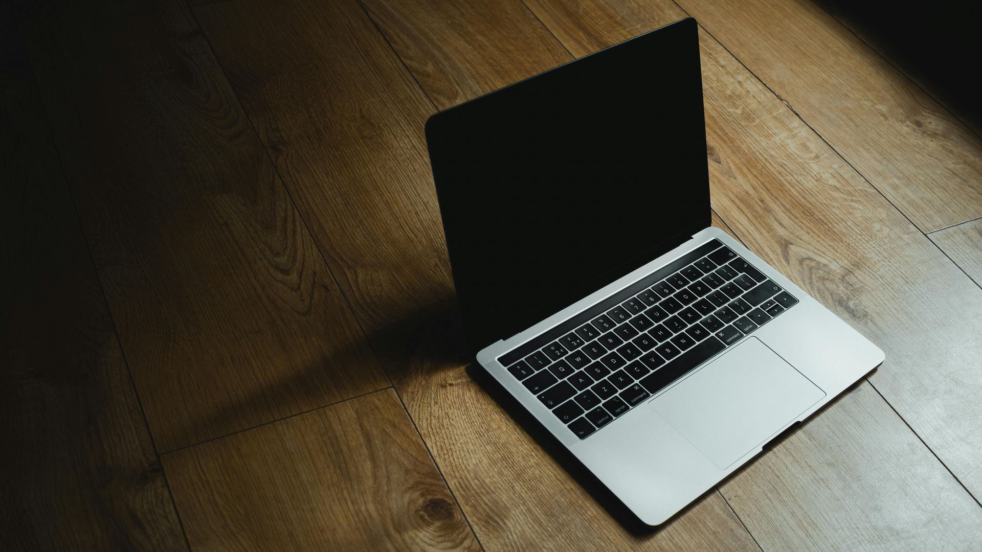 A silver laptop with a dark screen sitting on a wooden floor in dim, dramatic lighting.