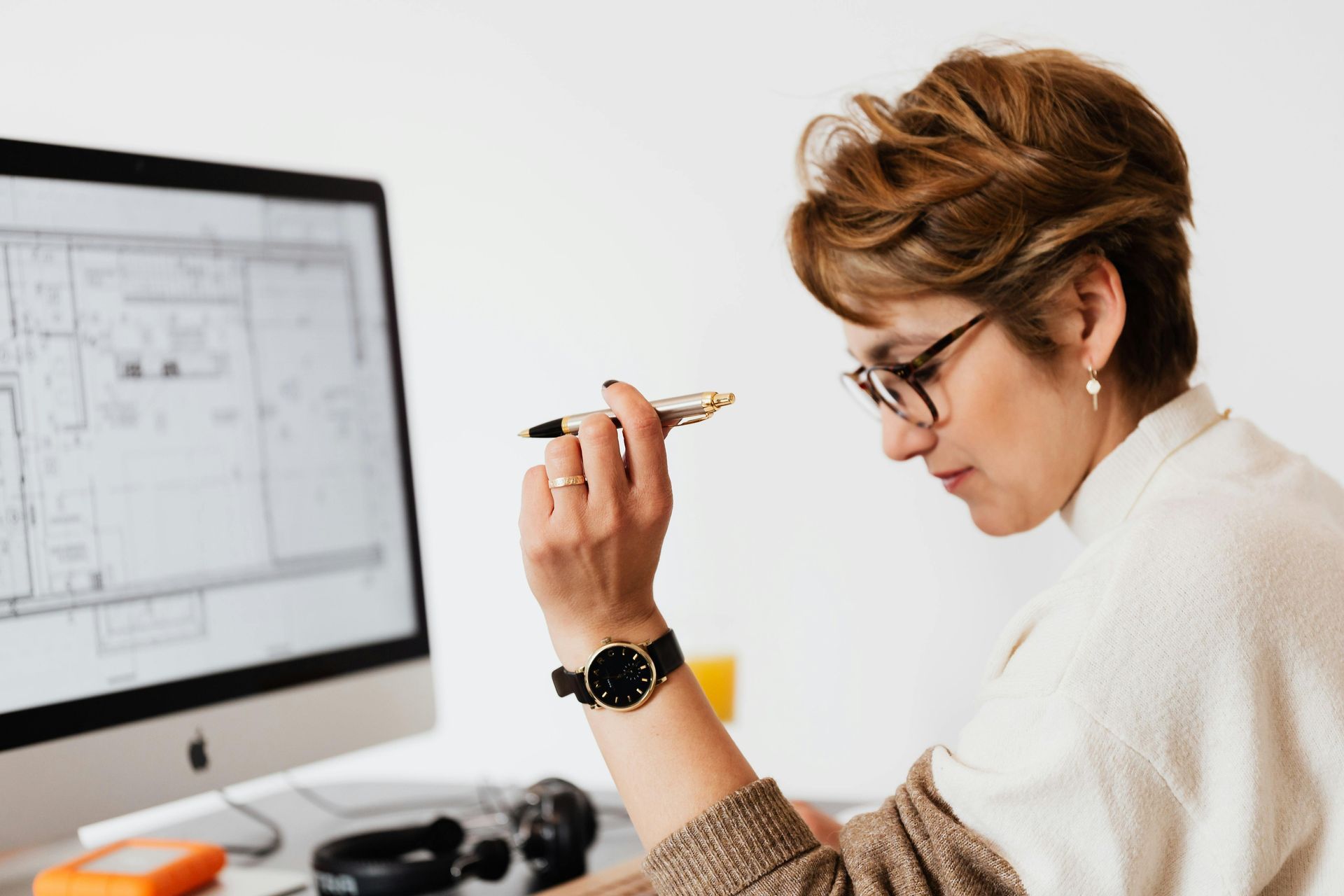 Woman with glasses looking at a computer screen, holding a pen. She is wearing a watch, sitting at a desk.
