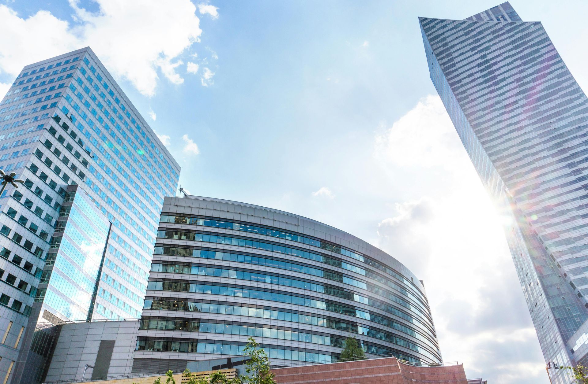 Low-angle view of three modern glass skyscrapers against a bright, sunny sky with scattered white clouds.