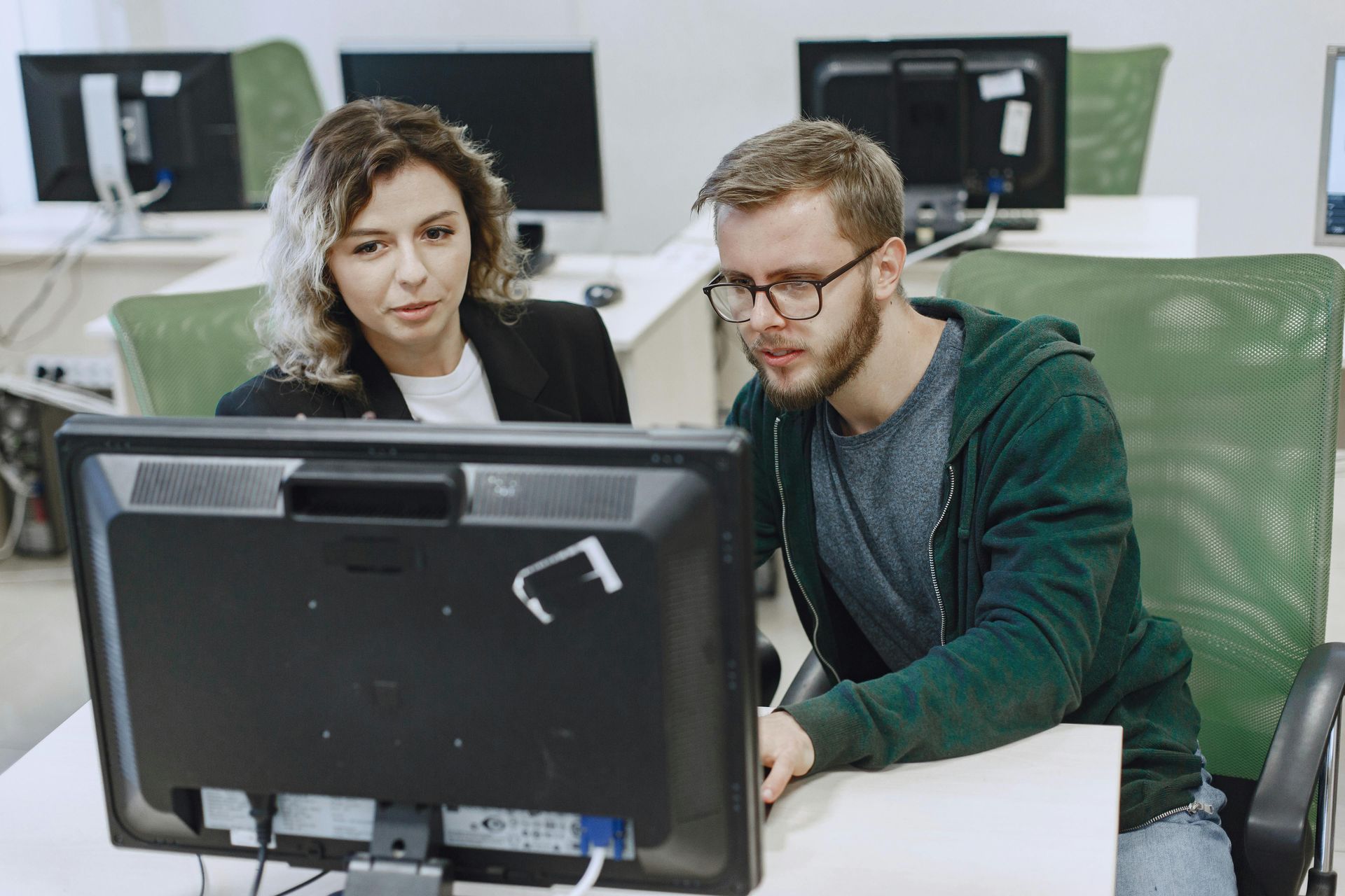 Two people seated at computer desks in an office, looking at a computer screen while collaborating on a project.