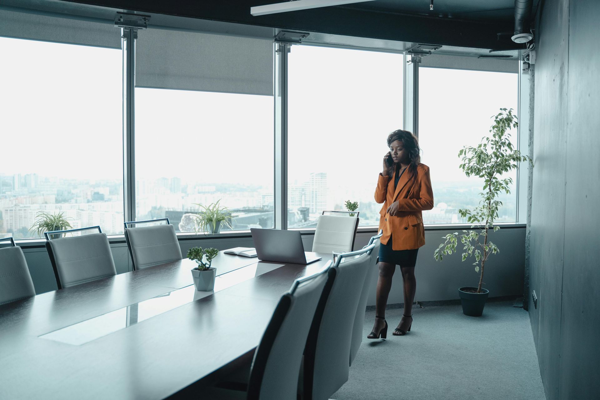 A person in a tan blazer talks on a phone while standing in a modern office boardroom overlooking a city.