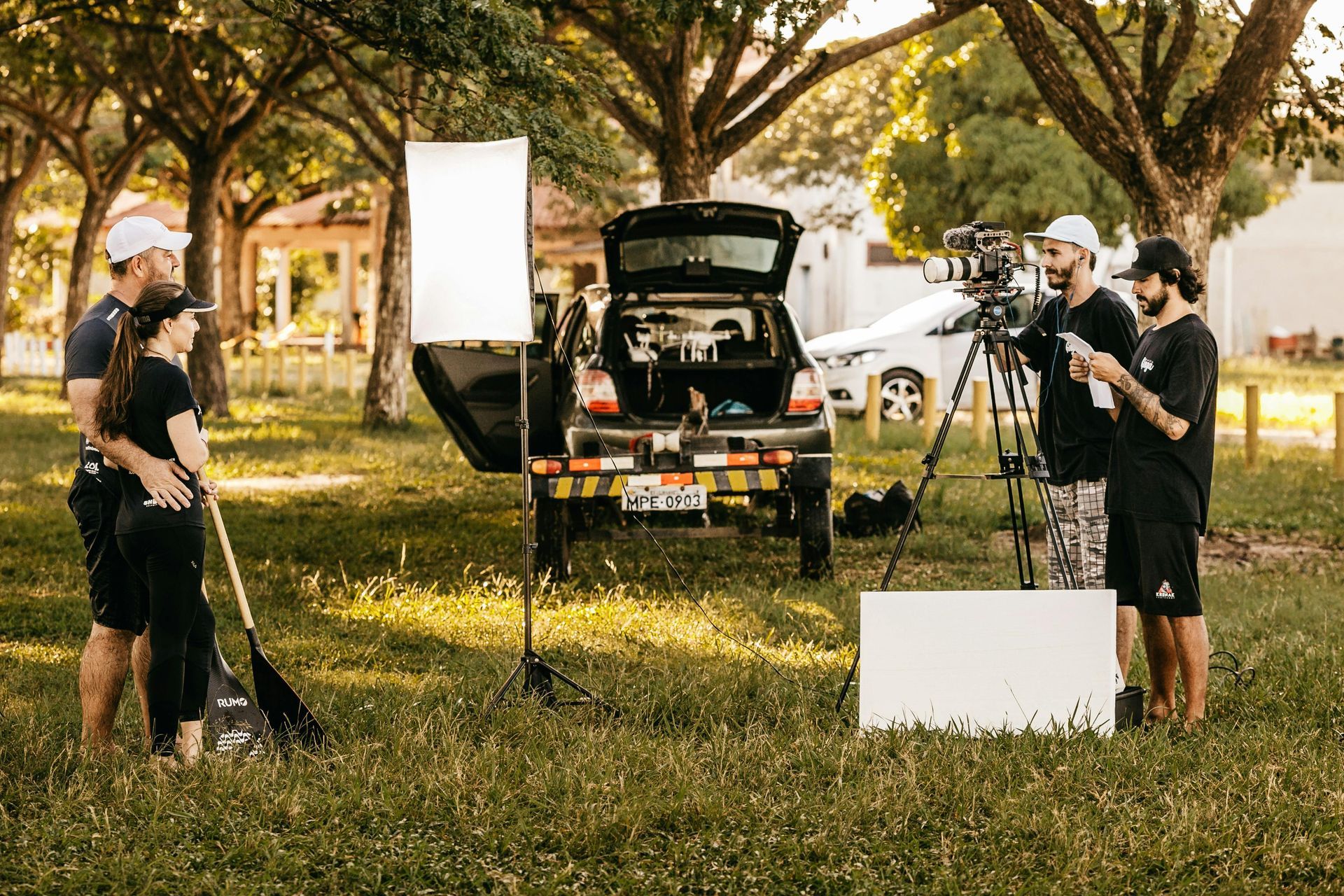 A video crew filming two people with a paddle in a grassy park near a car with open trunk and photography lighting gear.