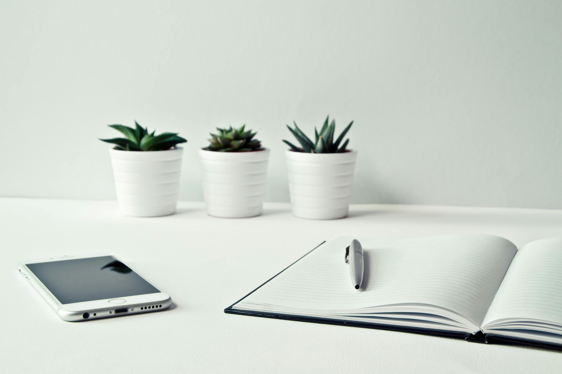 White desk with three succulents, a smartphone, a notebook, and a pen.
