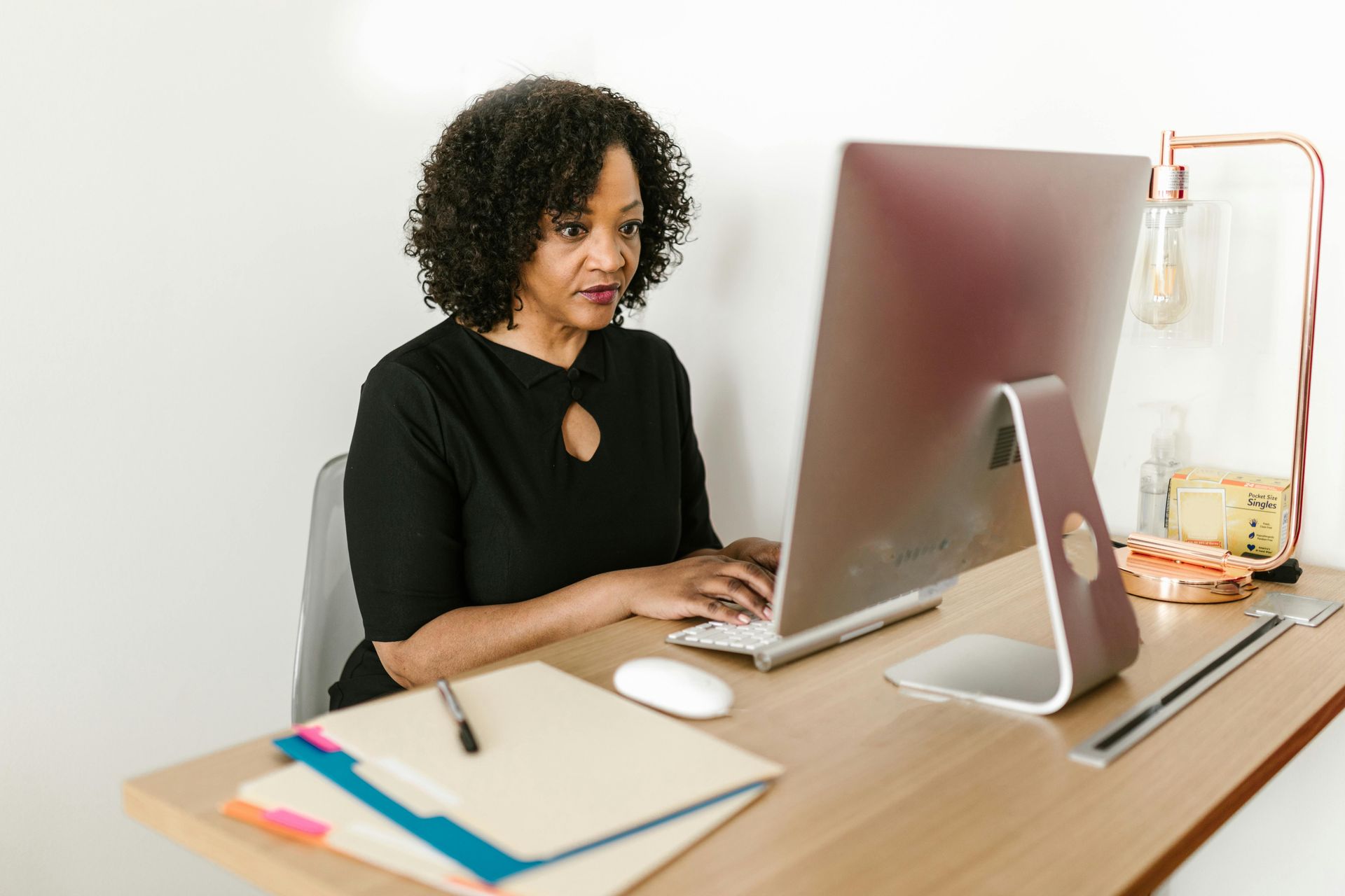 A person with dark curly hair wearing a black shirt works at a computer on a wooden desk in a bright office.