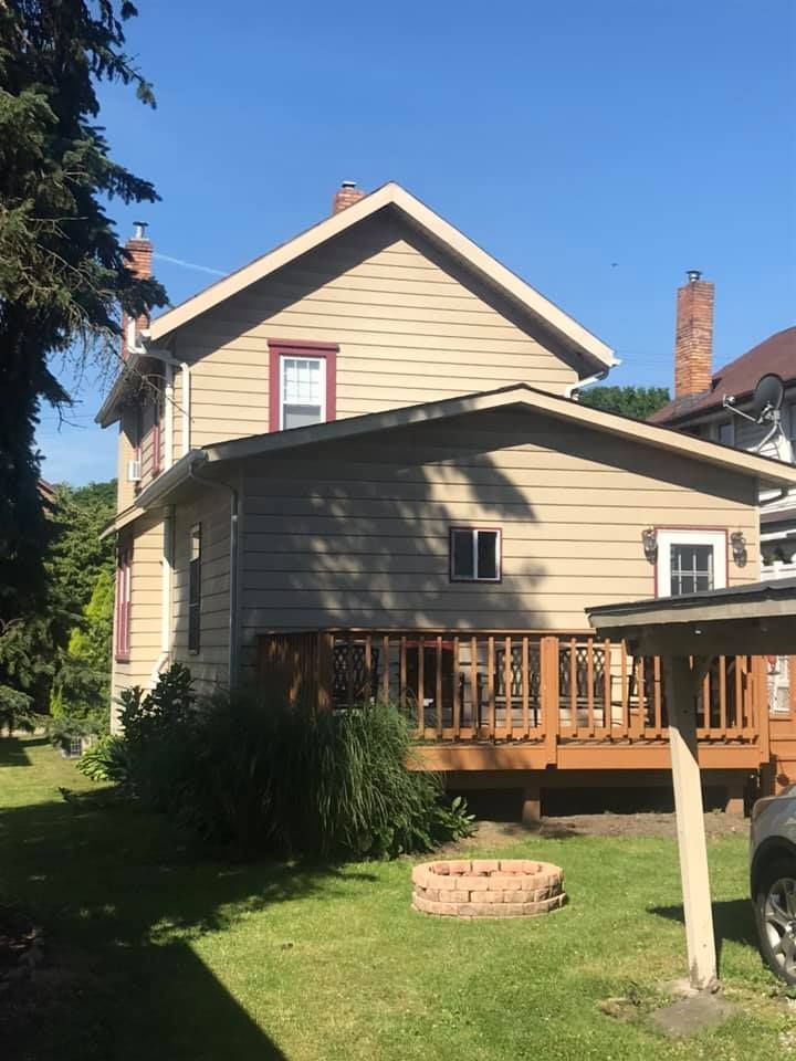 Tan two-story house with a wooden deck, a brick fire pit, and a green lawn under a blue sky.