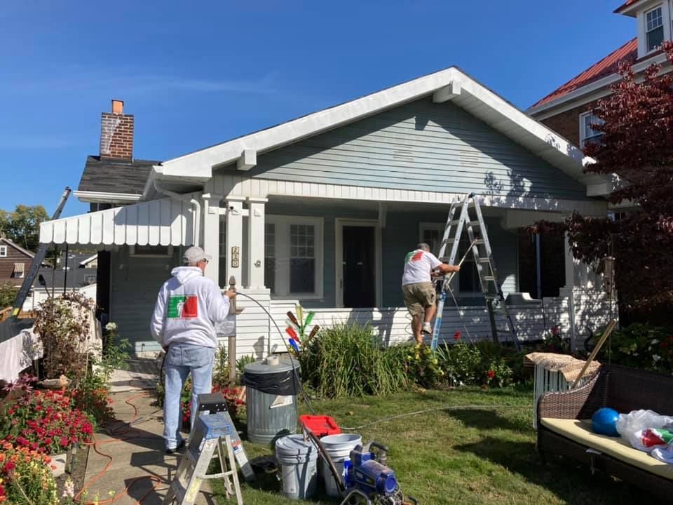 Two people painting a light blue house on a sunny day. One on a ladder, the other standing near the porch.