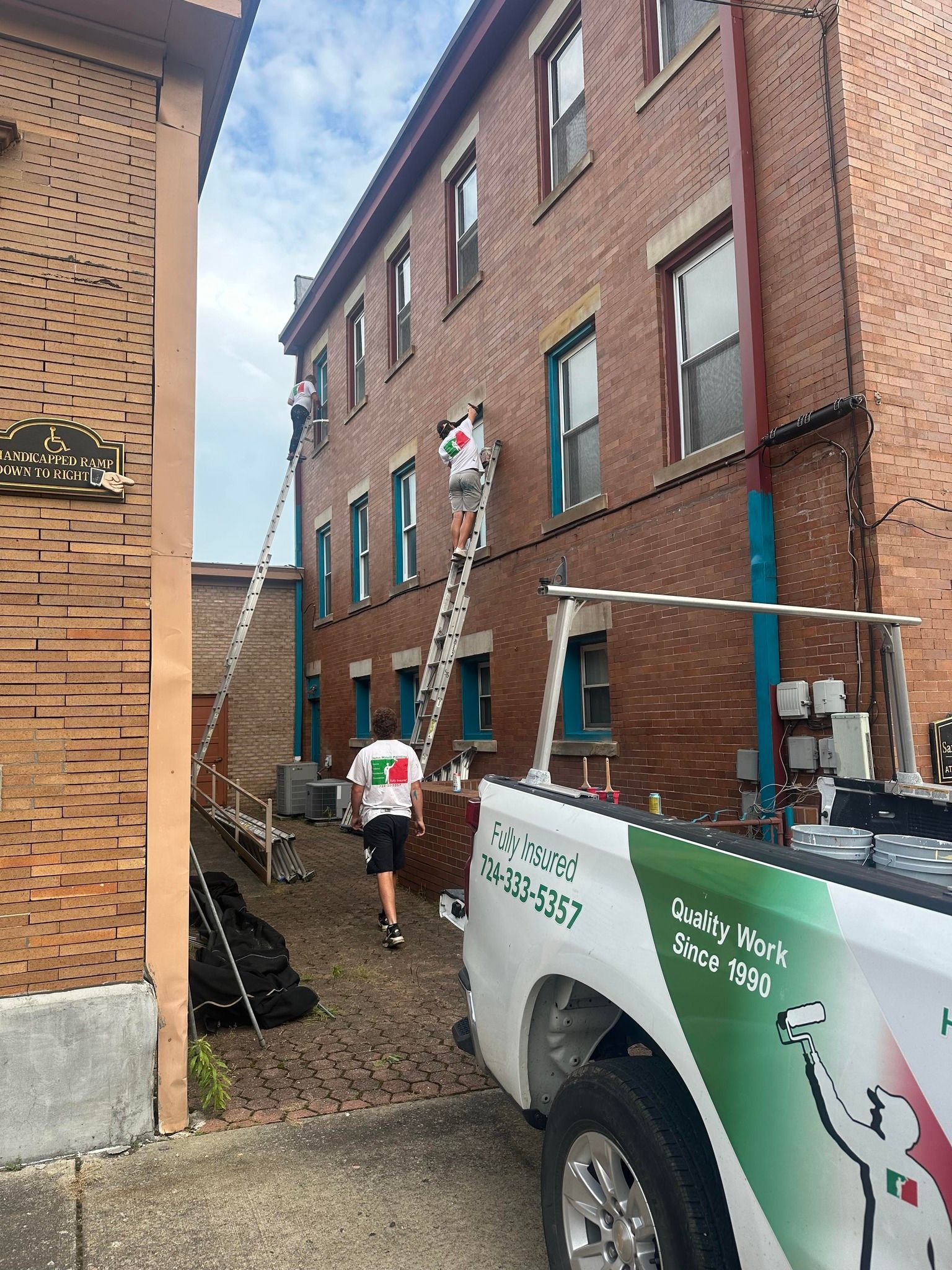 Workers on ladders near a brick building. Truck with logo. Cloudy sky.