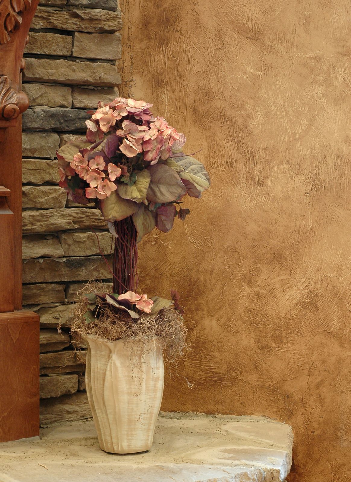 Plant with pink flowers in a light-colored pot on a stone step, against a textured wall.