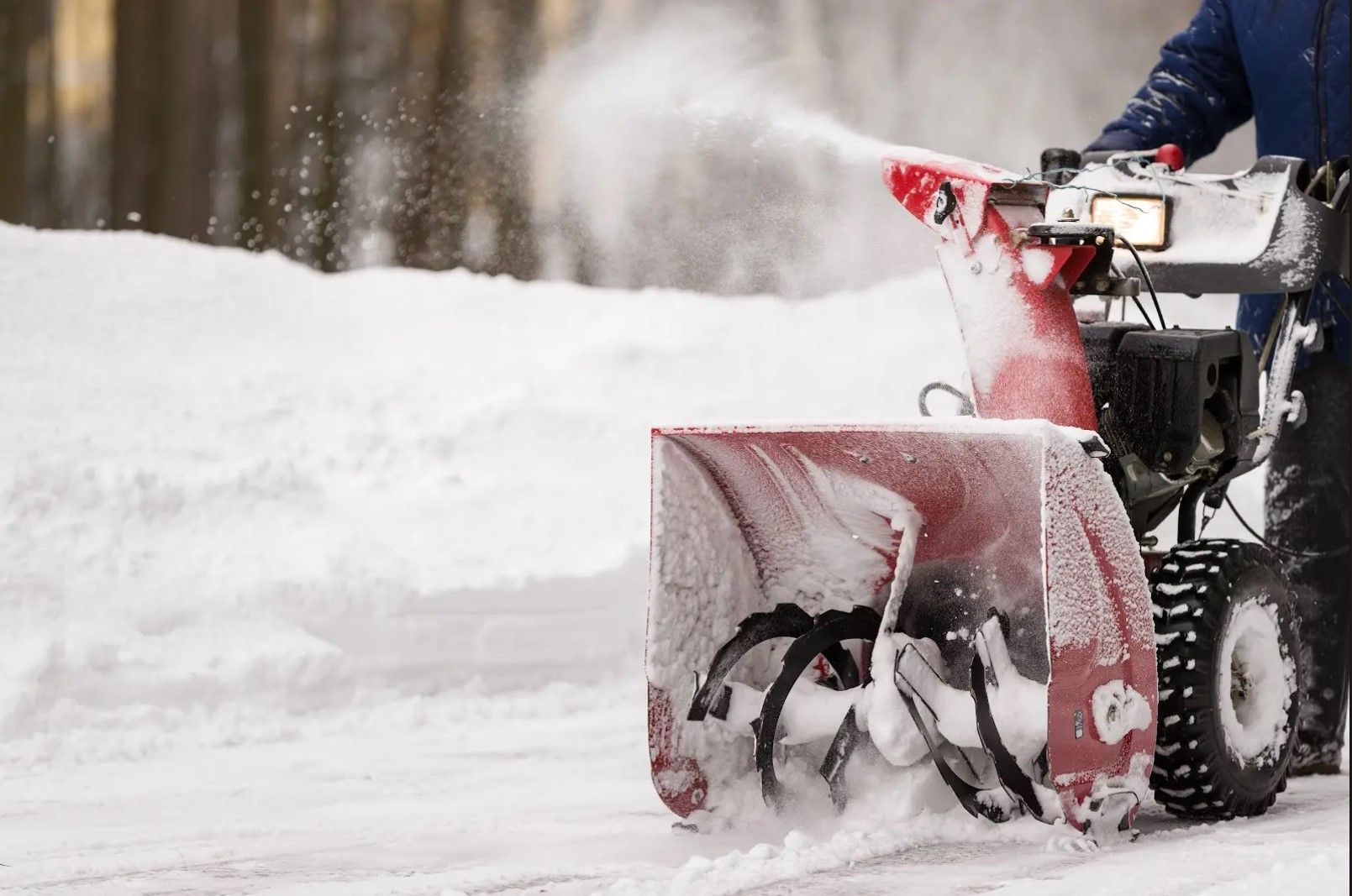 A person using a red snowblower to clear snow from a paved surface in a snowy outdoor setting.
