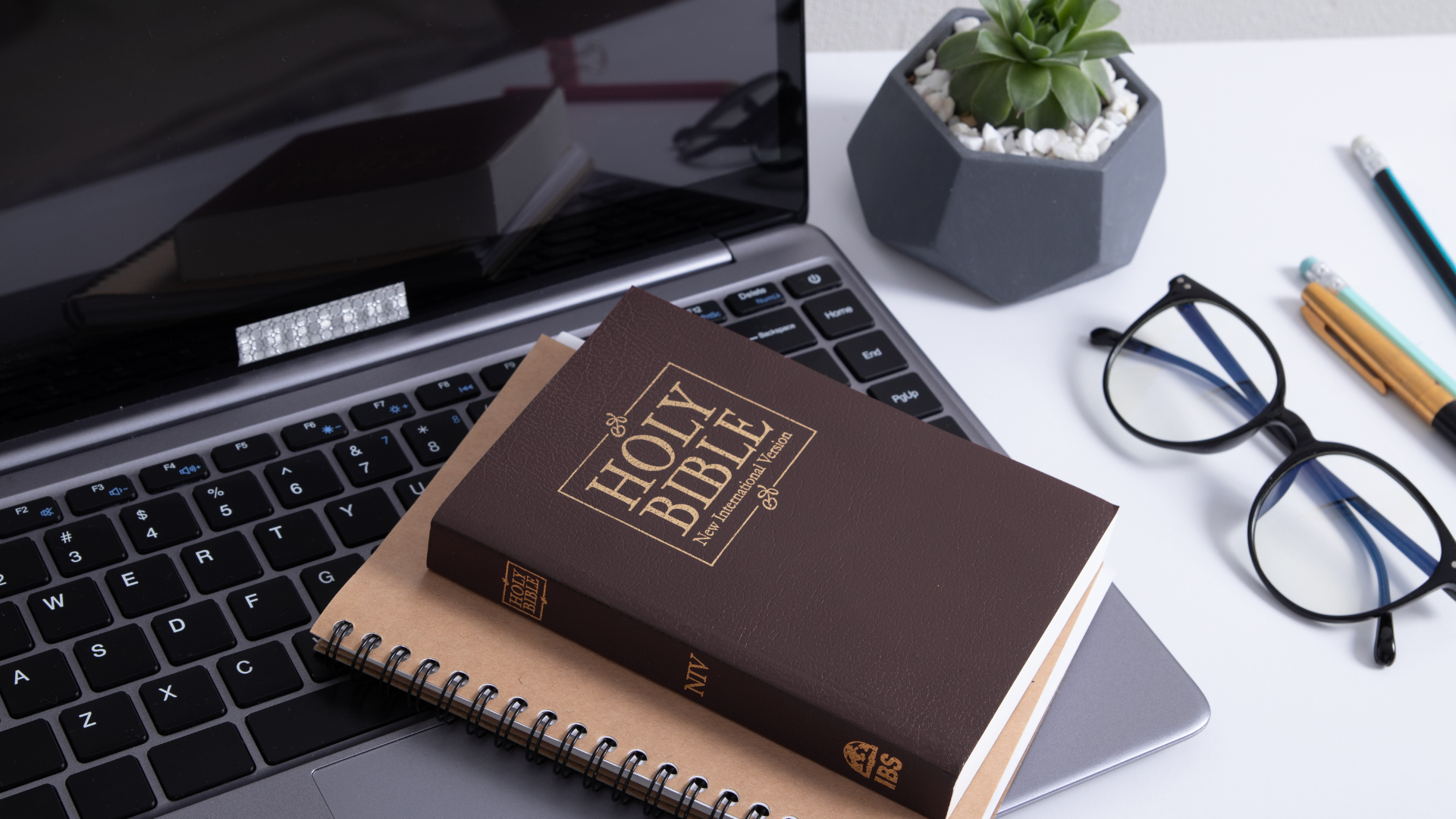 A holy bible is sitting on top of a notebook next to a laptop.