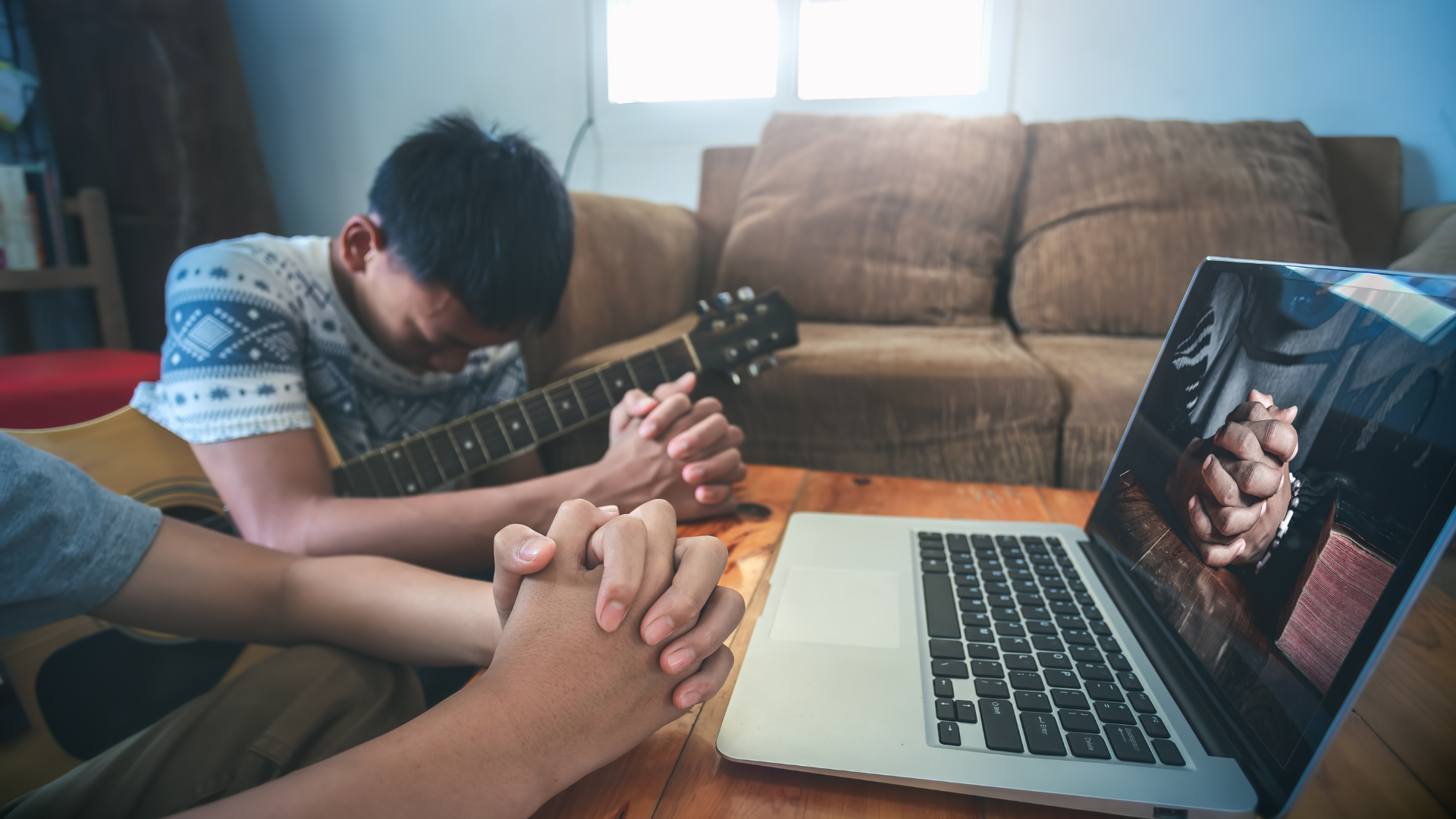 A man is playing a guitar while another man prays in front of a laptop computer.