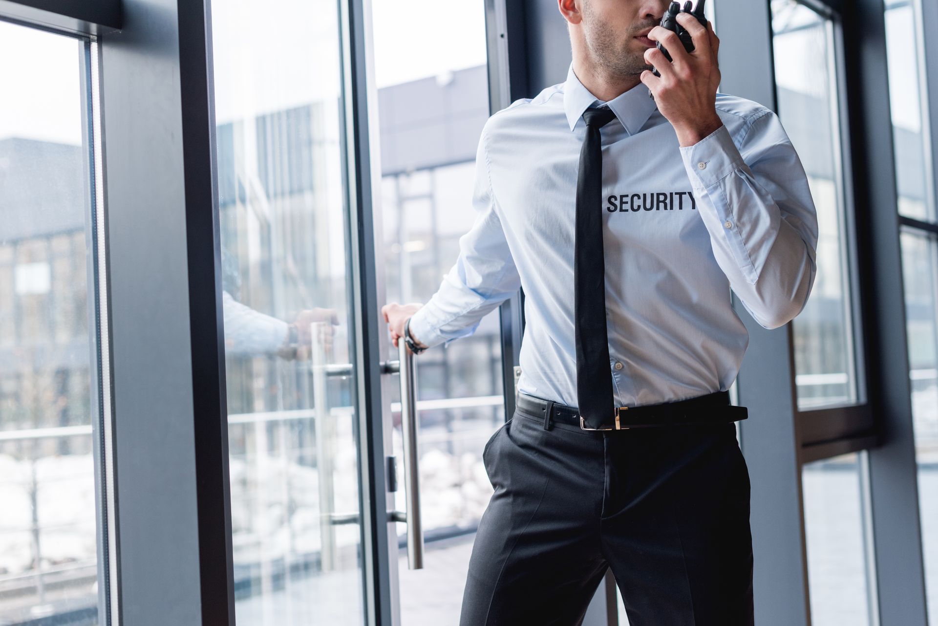 Security guard in a white shirt and black tie speaking into a radio by a glass office entrance