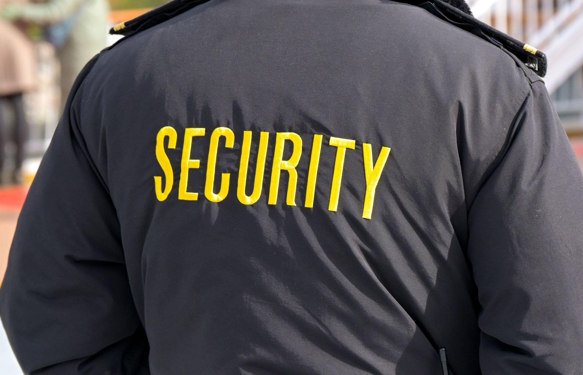 Back view of a security guard in uniform with “SECURITY” printed in bold yellow letters.