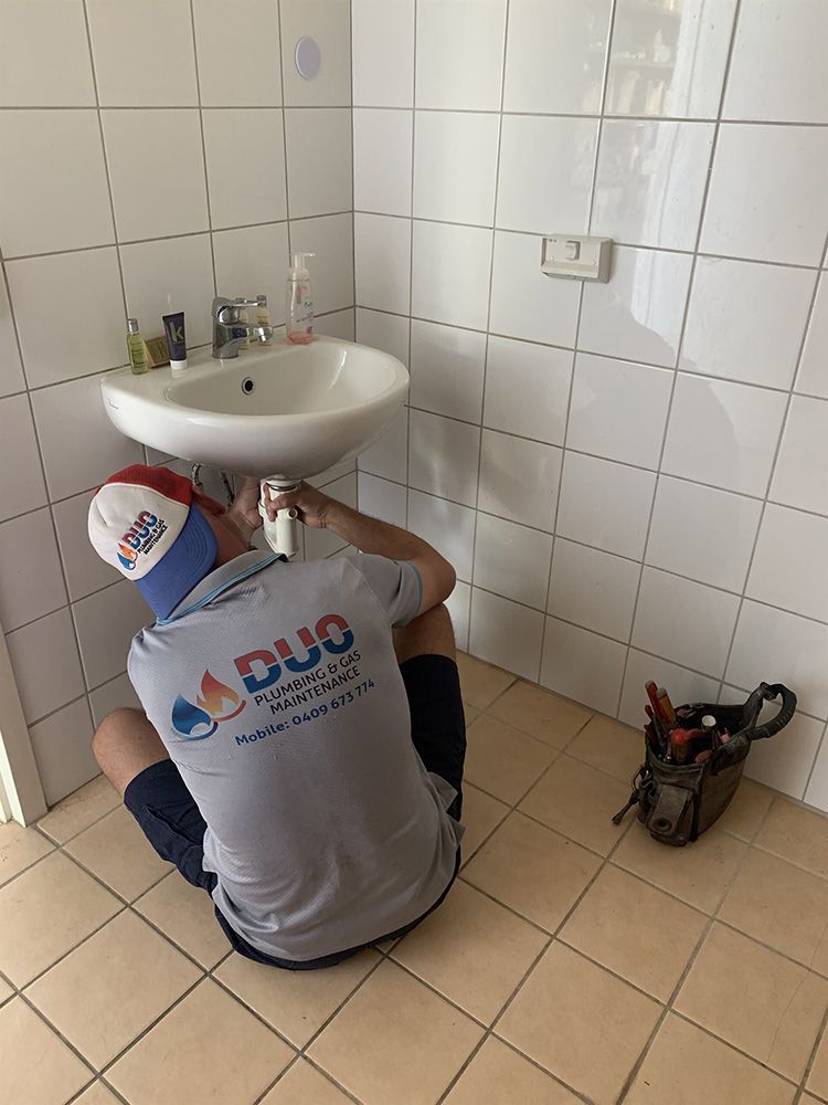 A Man is Kneeling Under a Sink in a Bathroom — Plumbers & Gas Fitters in Coconut Grove, NT