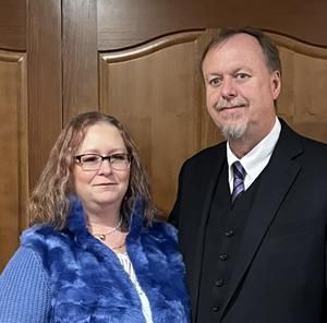 Woman in blue vest and man in suit, smiling in front of wood paneling.