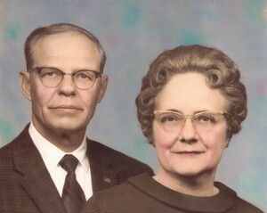 Man and woman in glasses, formal wear; posing for a portrait against a mottled background.
