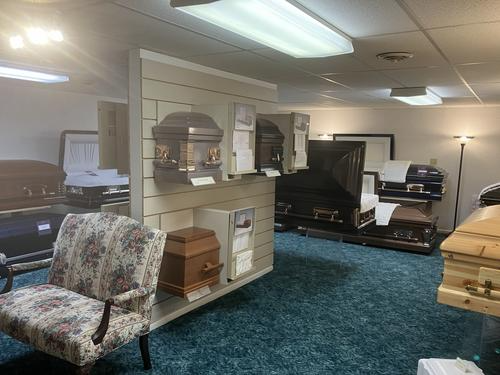 Interior of a funeral home displaying various caskets, a patterned chair, and teal carpet.
