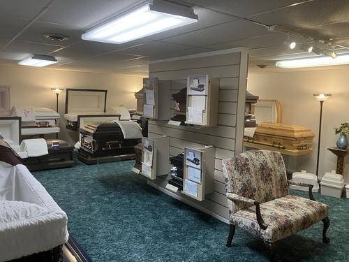 Funeral home showroom with caskets on display and a patterned armchair on a teal carpet.