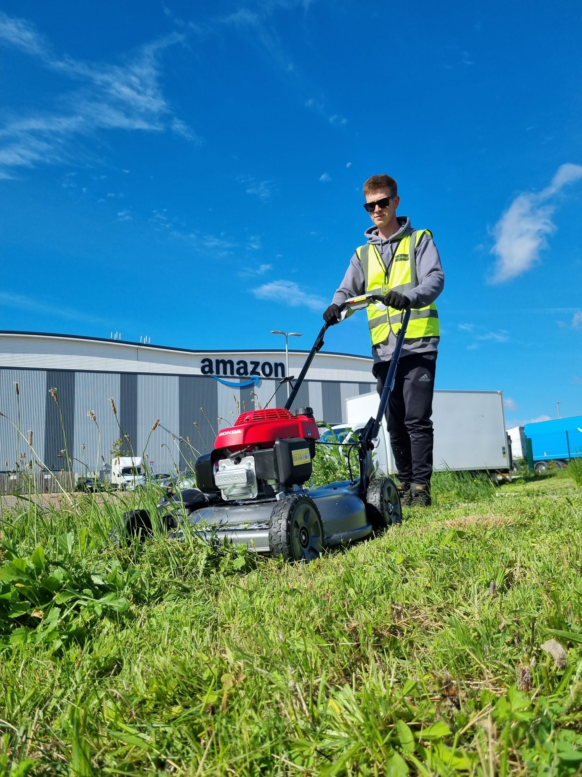 A man is cutting grass with a lawn mower in front of an amazon building.
