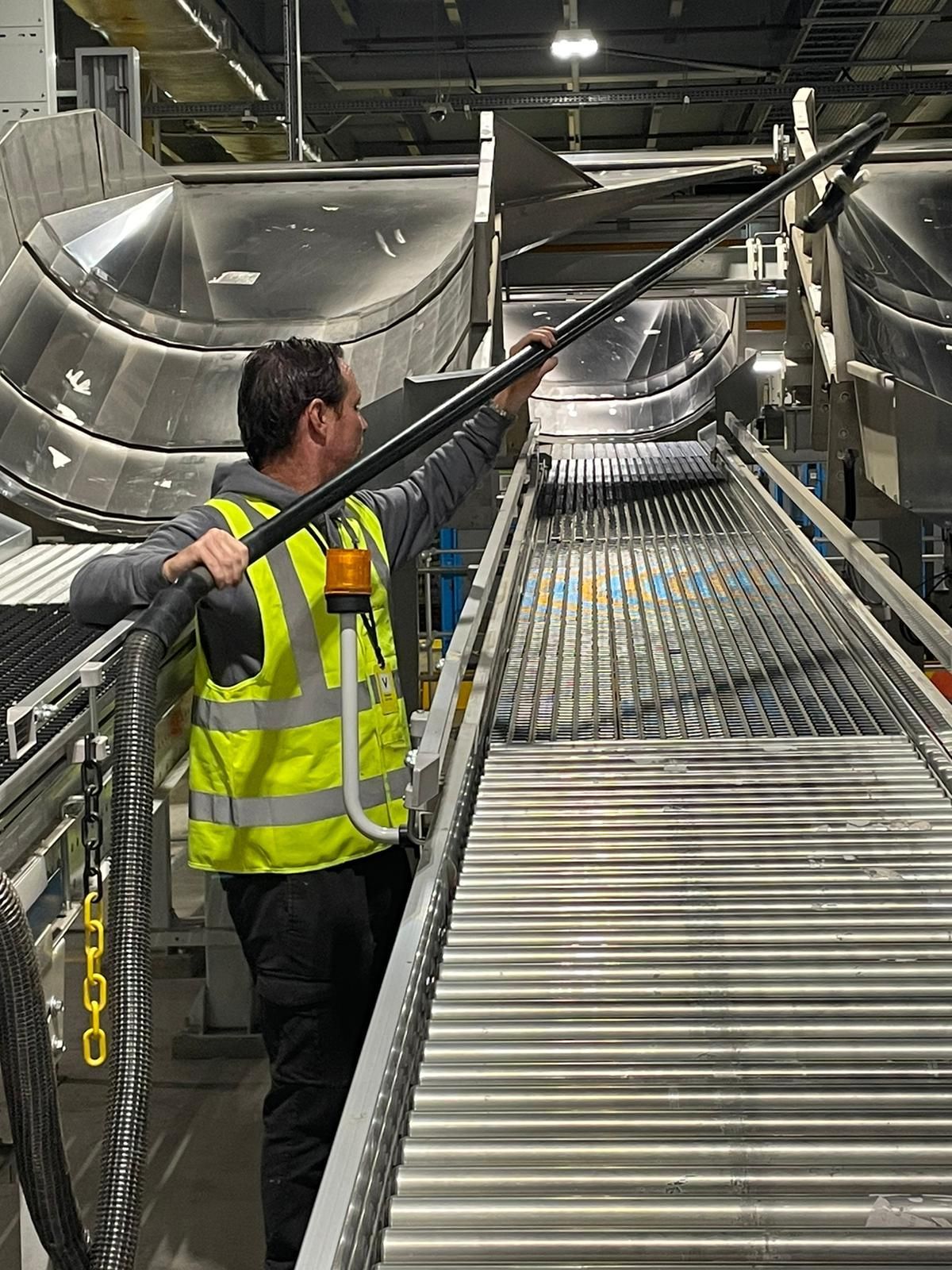 A man in a yellow vest is cleaning a conveyor belt with a vacuum cleaner.
