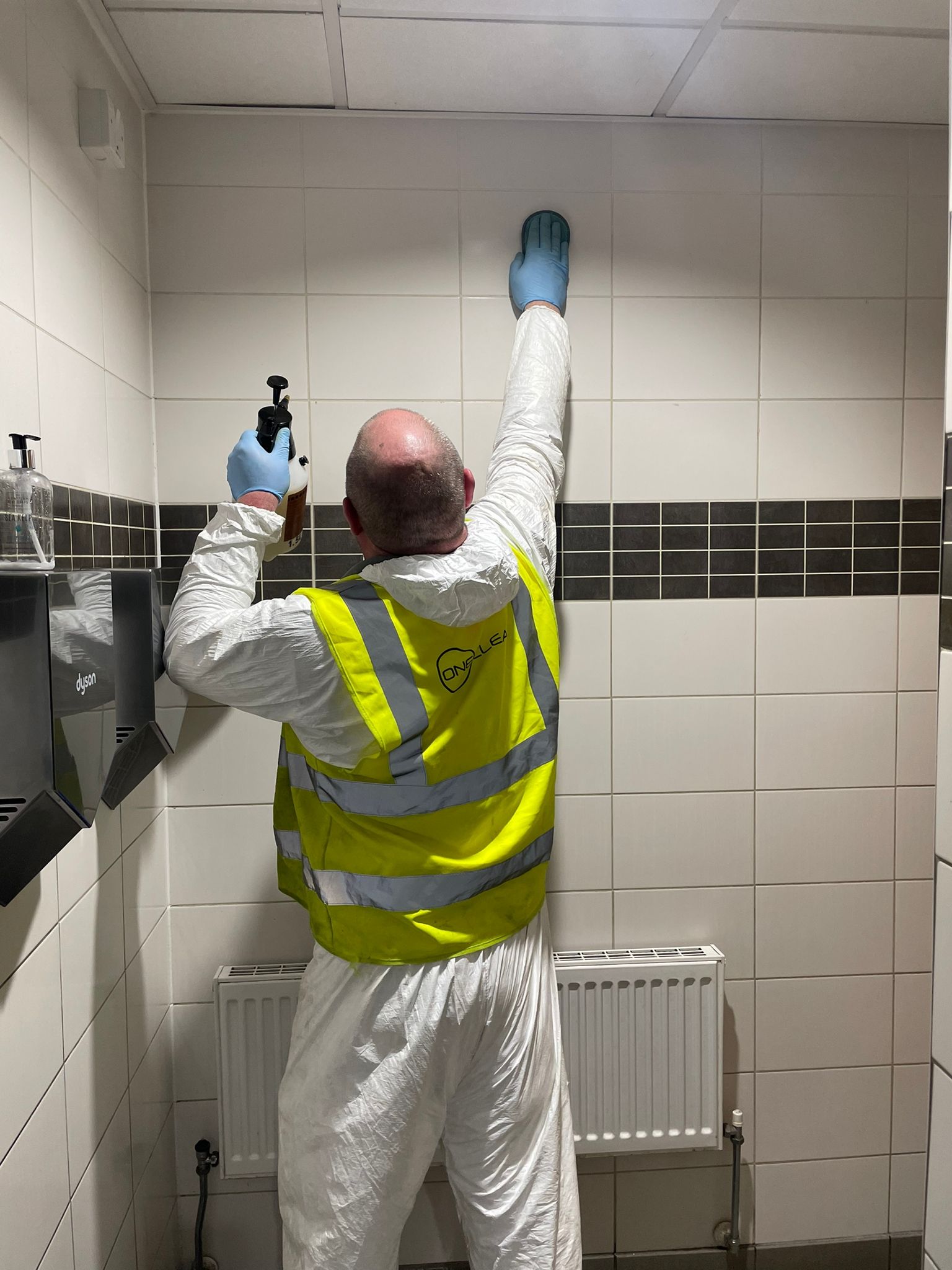 A man in a yellow vest is cleaning a wall in a bathroom.
