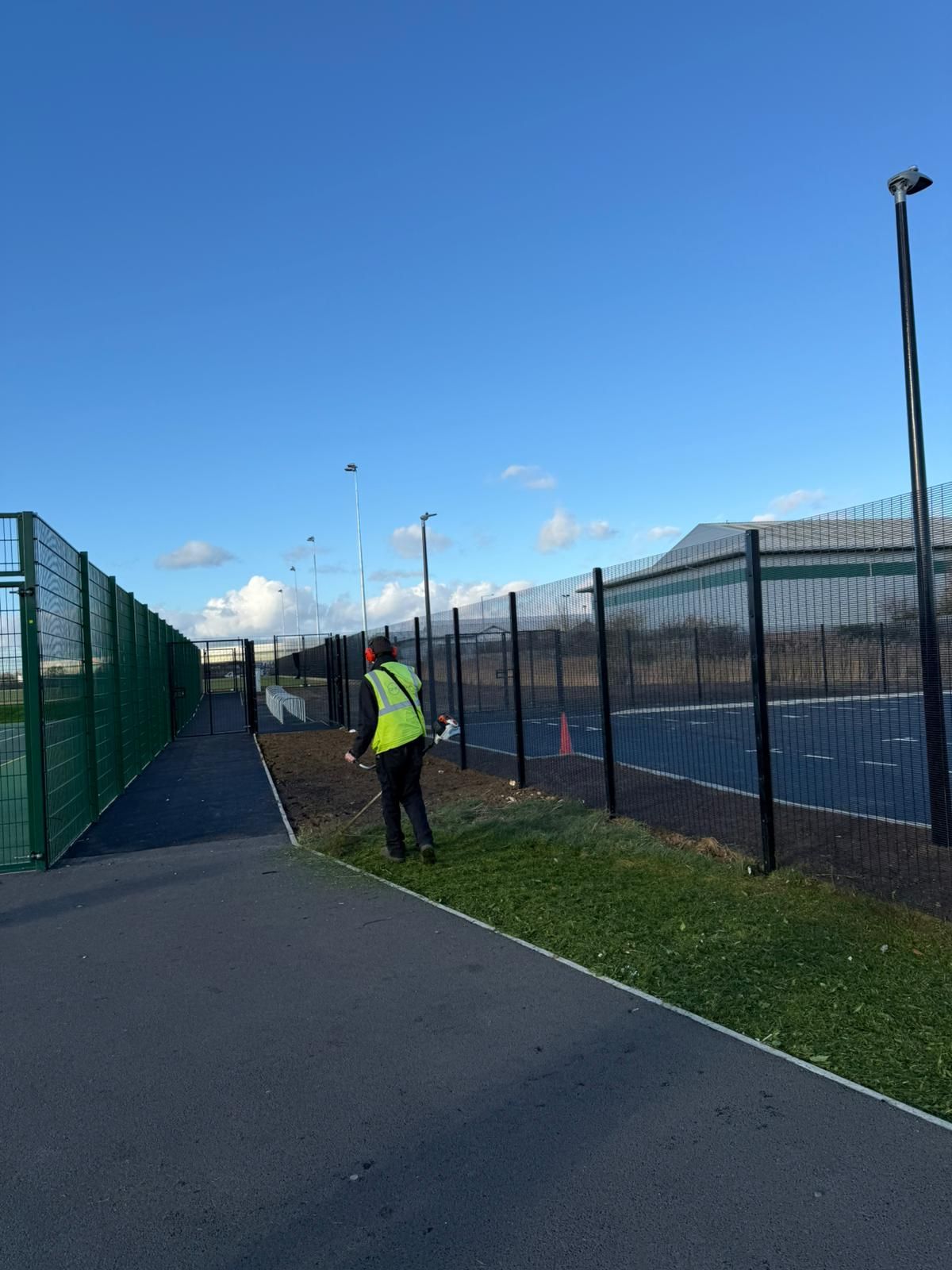 A man in a yellow vest is walking down a path next to a tennis court.