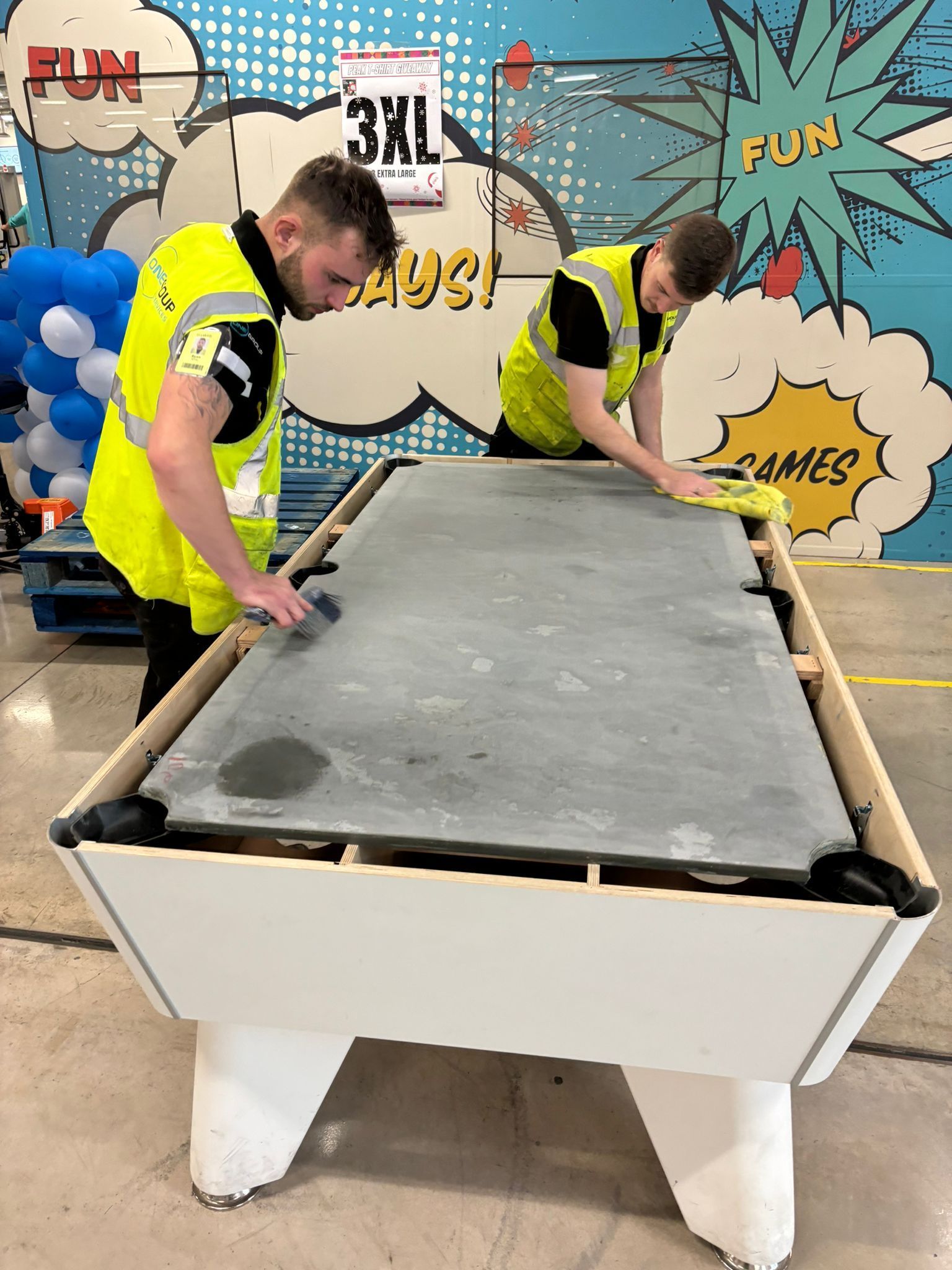 Two men in yellow vests are cleaning a pool table.