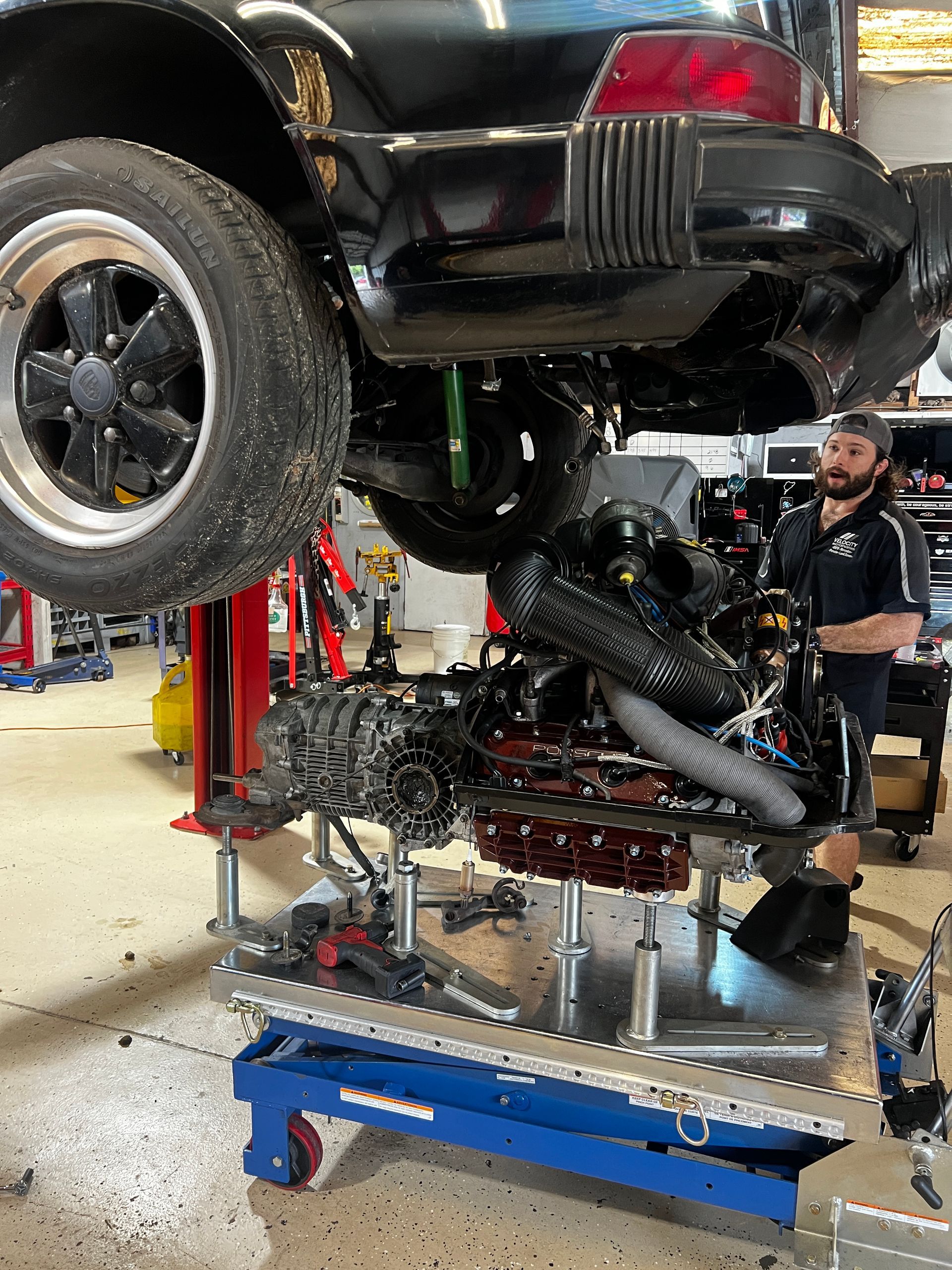 Mechanic working on a car; the engine is on a lift. The car is black, the engine is grey and red. | Velocity Motorwerks