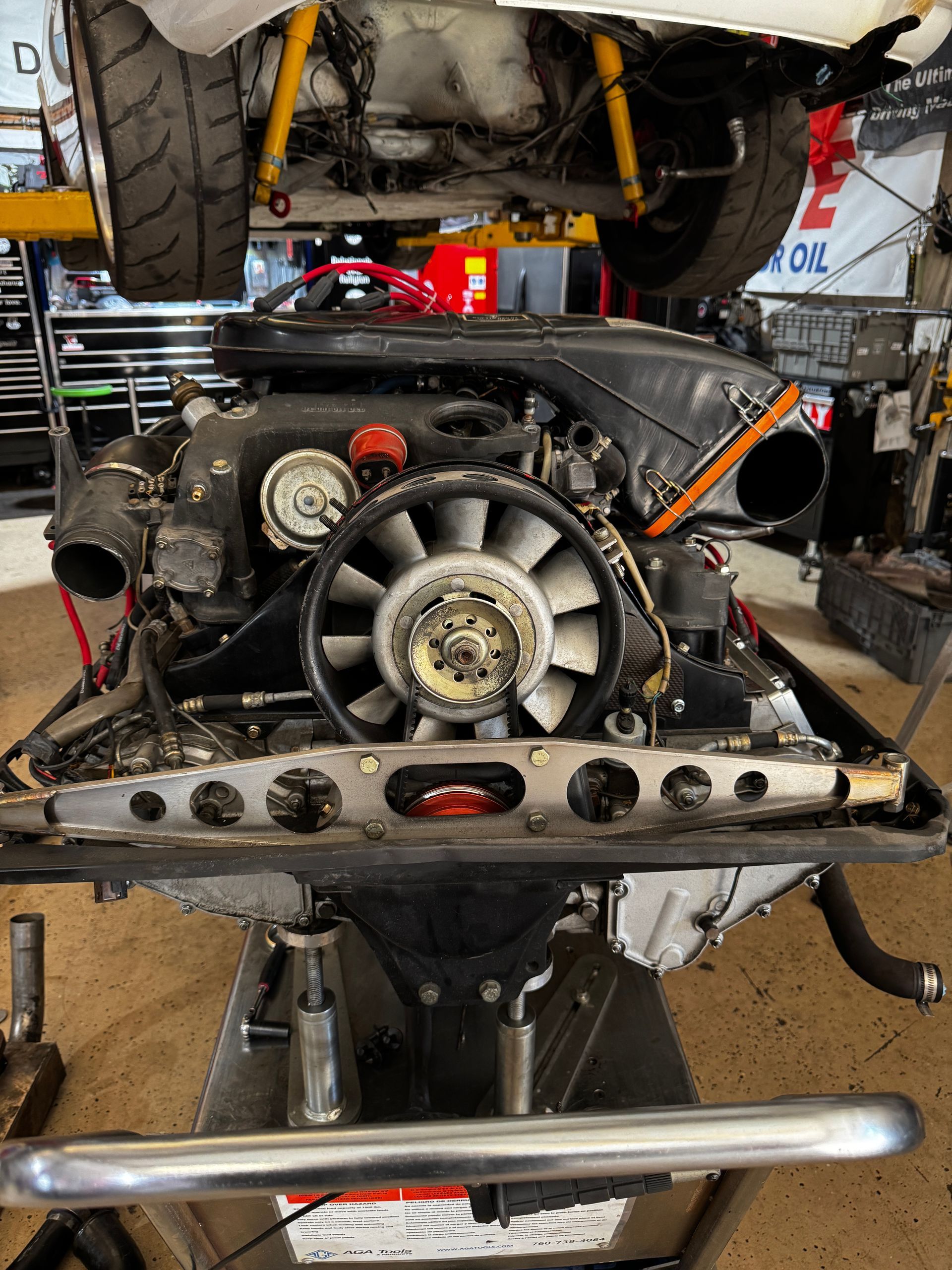 Porsche engine on a lift in a garage. The engine is black, with a silver fan and is in the process of being worked on. | Velocity Motorwerks