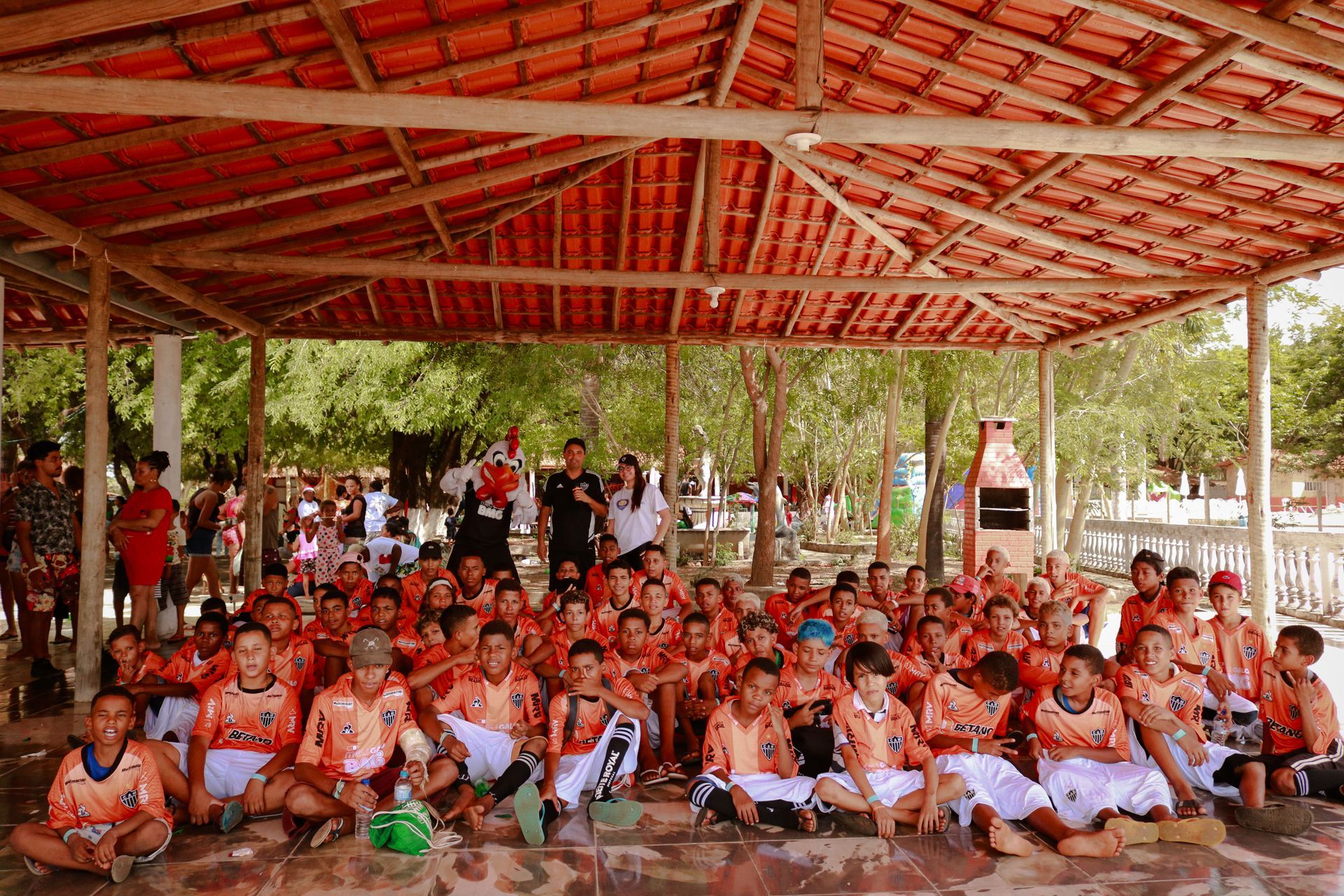 Um grande grupo de crianças está sentado sob uma estrutura de madeira.