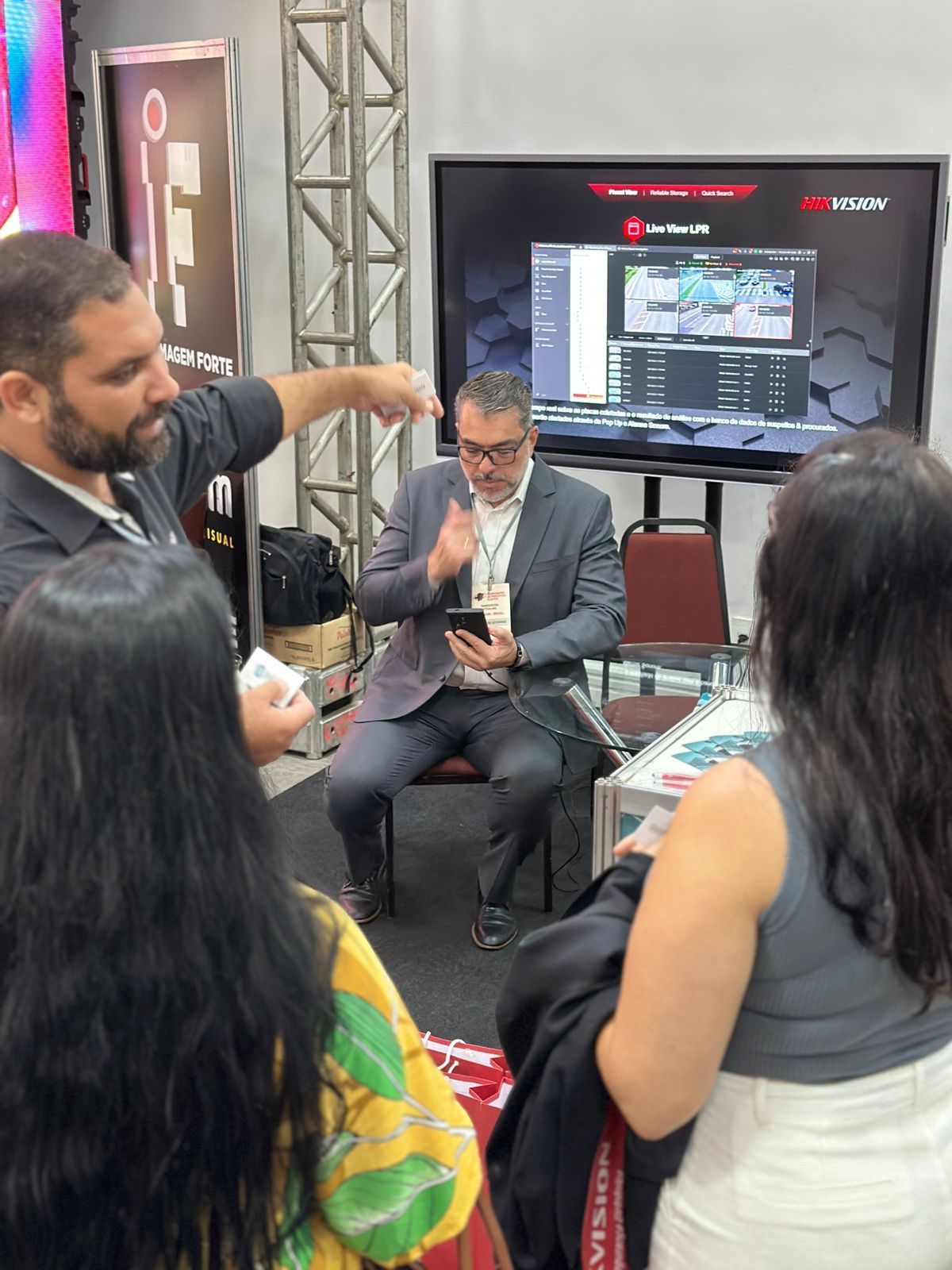 Um homem de terno está conversando com um grupo de pessoas em frente a uma tela grande.