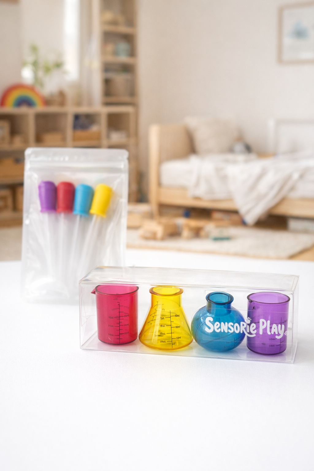 Colorful plastic laboratory beakers and droppers from Sensorie Play on a wooden table in a brightly lit room.