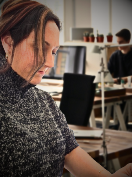 Une femme en pull gris, concentrée sur son travail, avec une personne et un bureau partiellement visibles en arrière-plan.