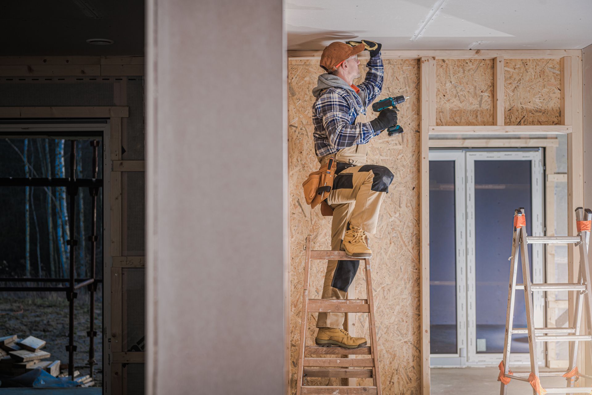 Worker installing drywall in a room, standing on a wooden ladder beside a window and scaffolding.
