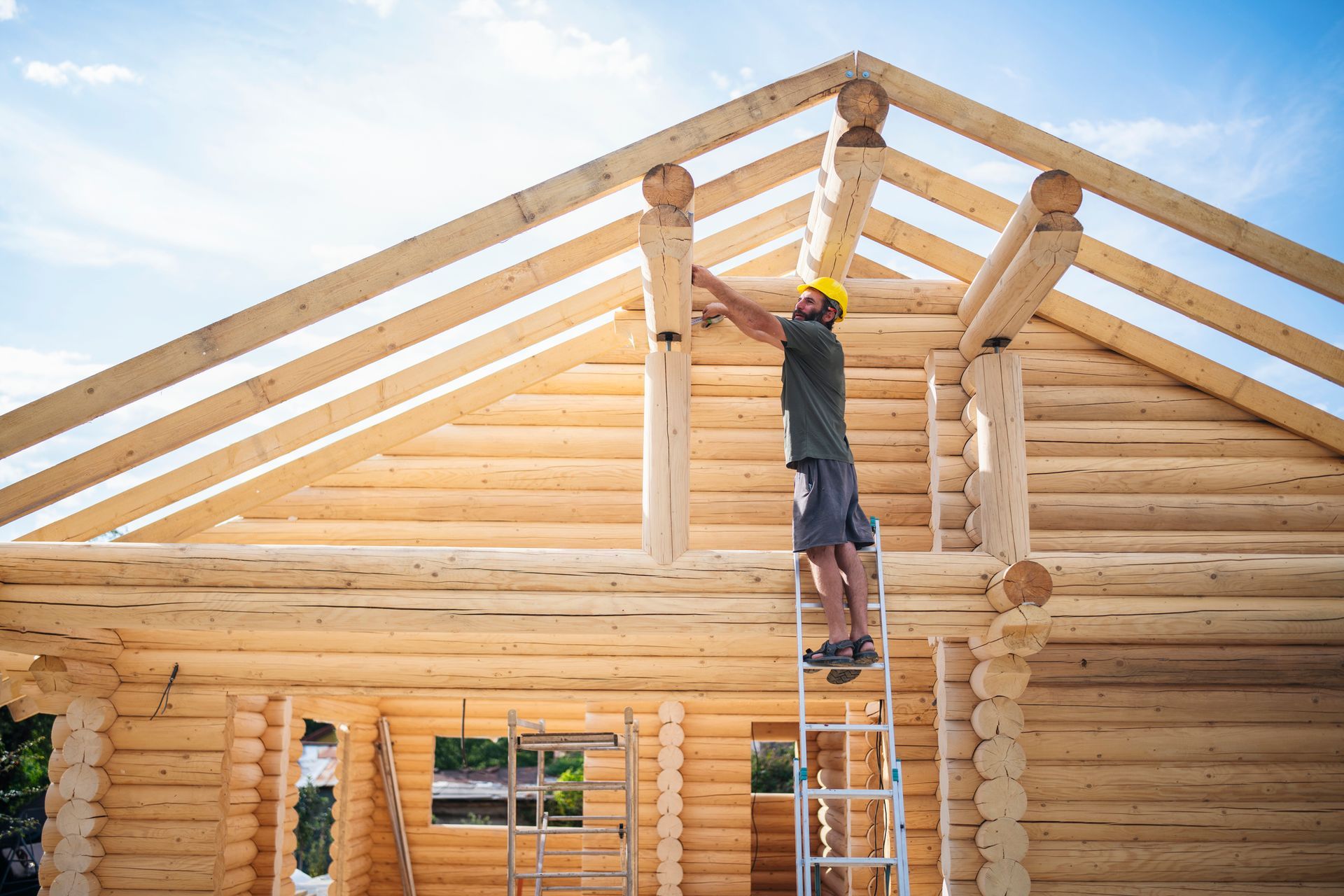 A man is standing on a ladder in front of a wooden house under construction.