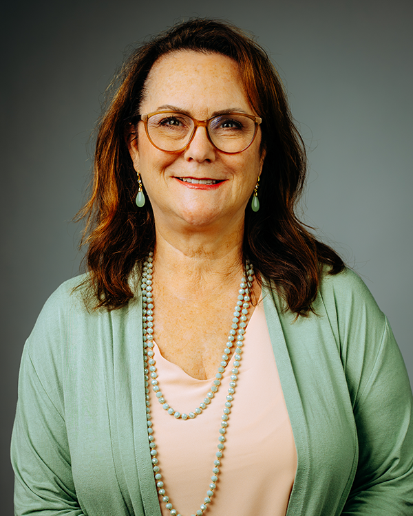 Woman with glasses and long hair smiles, wearing a black top and silver necklace against a dark background.