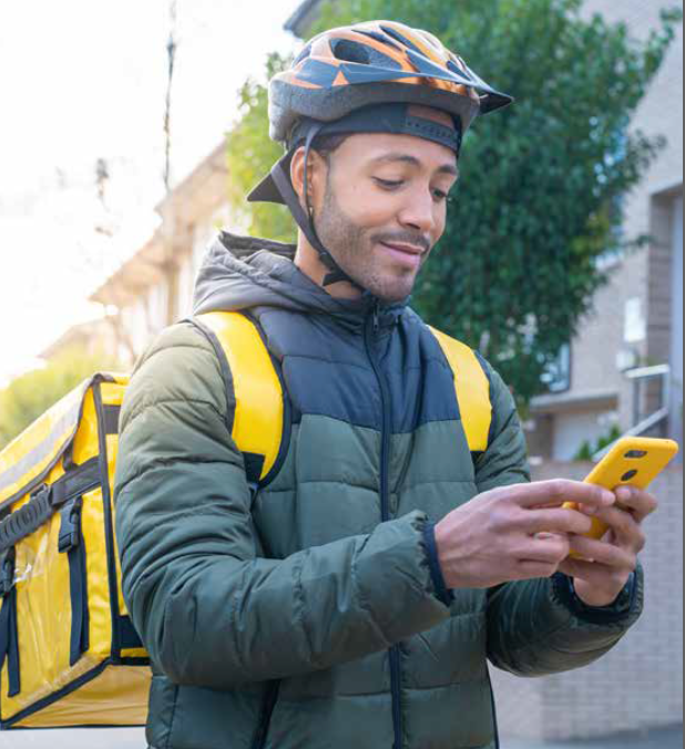 Man in helmet and delivery vest looking at phone, outdoors near building.