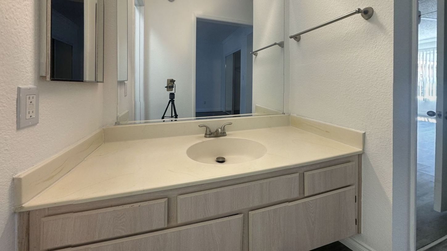 Bathroom with light-colored vanity, large mirror, and silver towel rack. A camera on a tripod is reflected in the mirror.