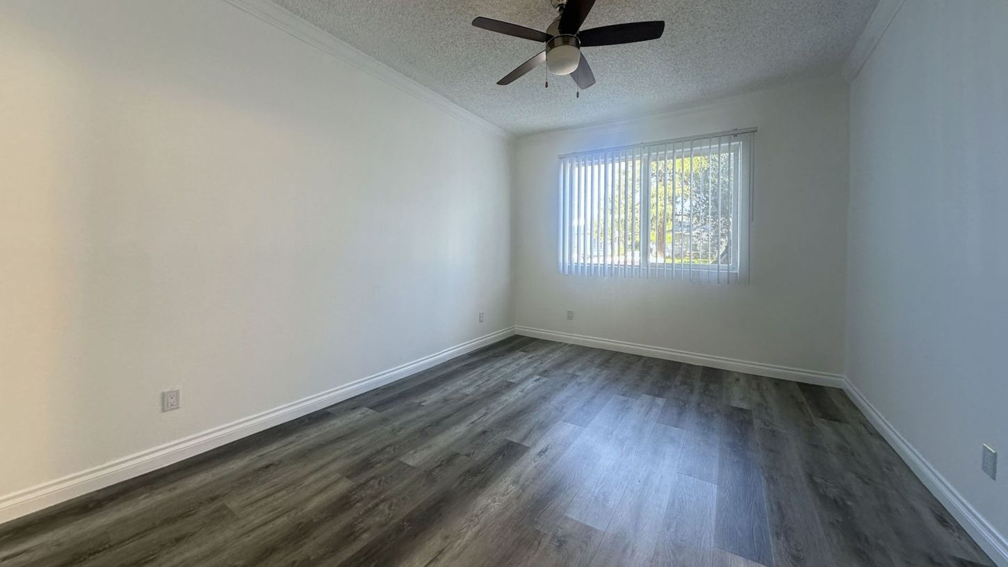 Empty room with gray wood-look floor, white walls, window with blinds, ceiling fan.