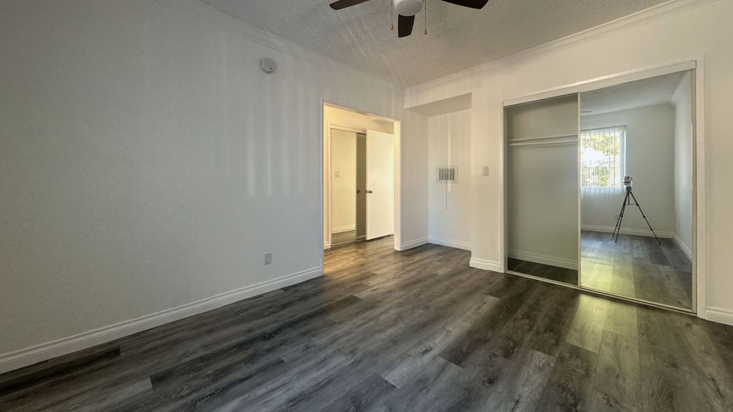 Empty bedroom with gray wood-look flooring, white walls, closet with mirrored doors, and an open doorway.