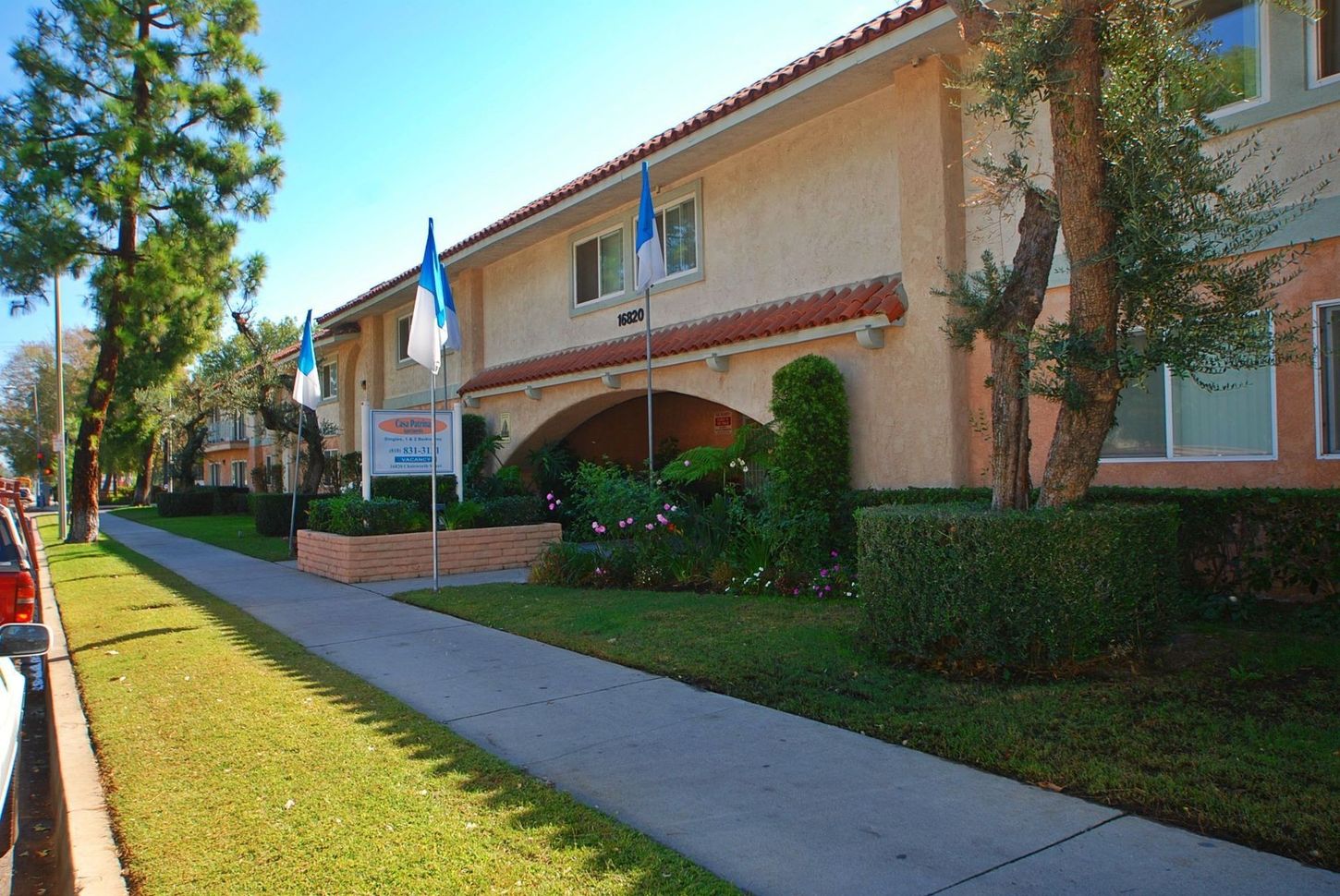 Apartment building with beige stucco, red tile roof, and blue flags. Green lawn and sidewalk in front.