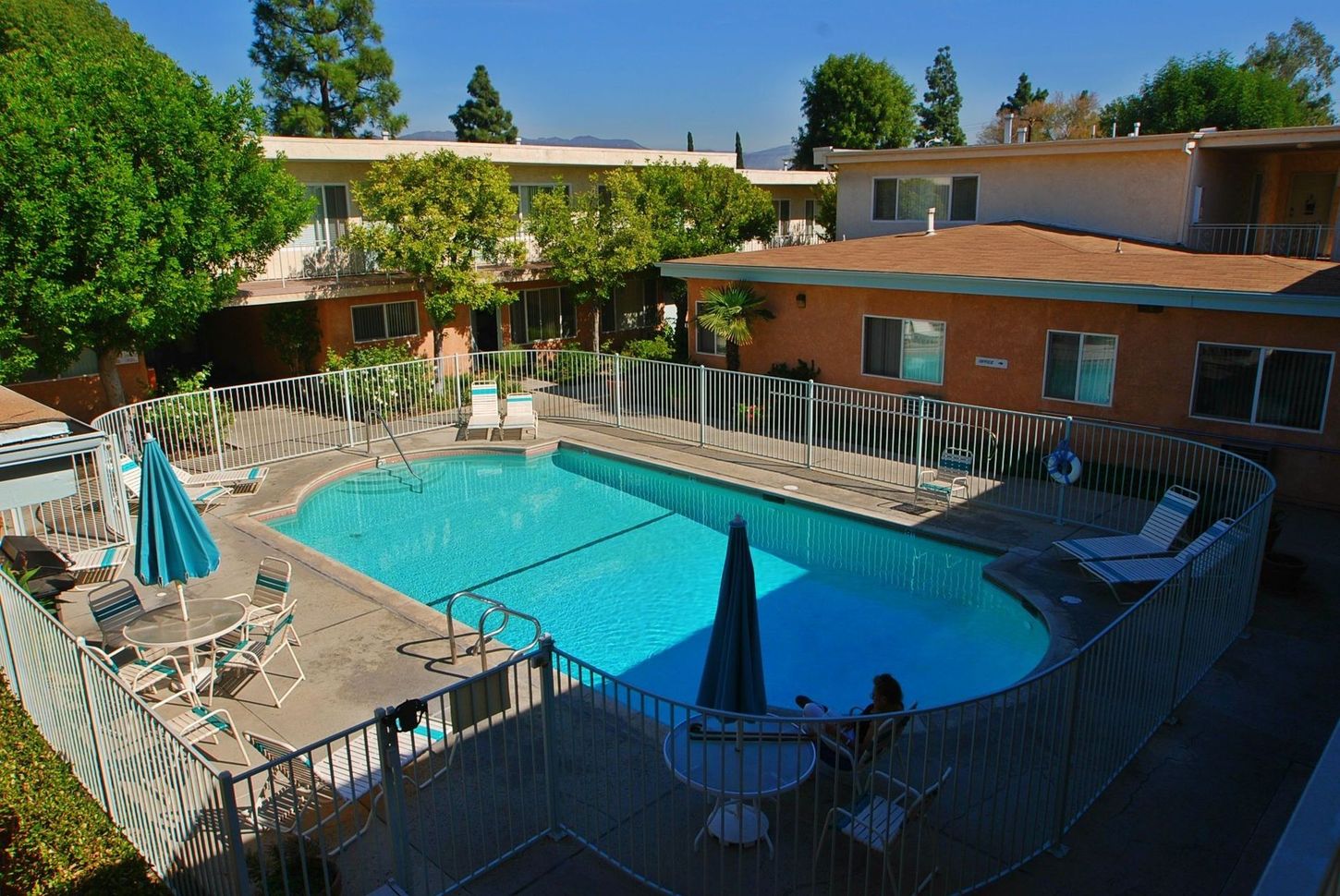 Pool surrounded by apartments, with blue water, fences, lounge chairs, and umbrellas.
