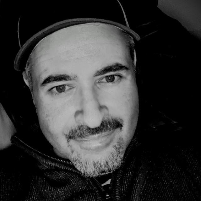 Man with graying hair and goatee wearing a baseball cap, smiling. Black and white.