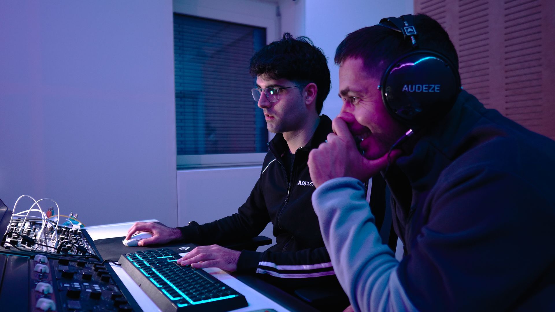 Zwei Personen mit Headsets arbeiten an einem Gaming-Setup und konzentrieren sich auf Bildschirme in violett-blauer Beleuchtung.
