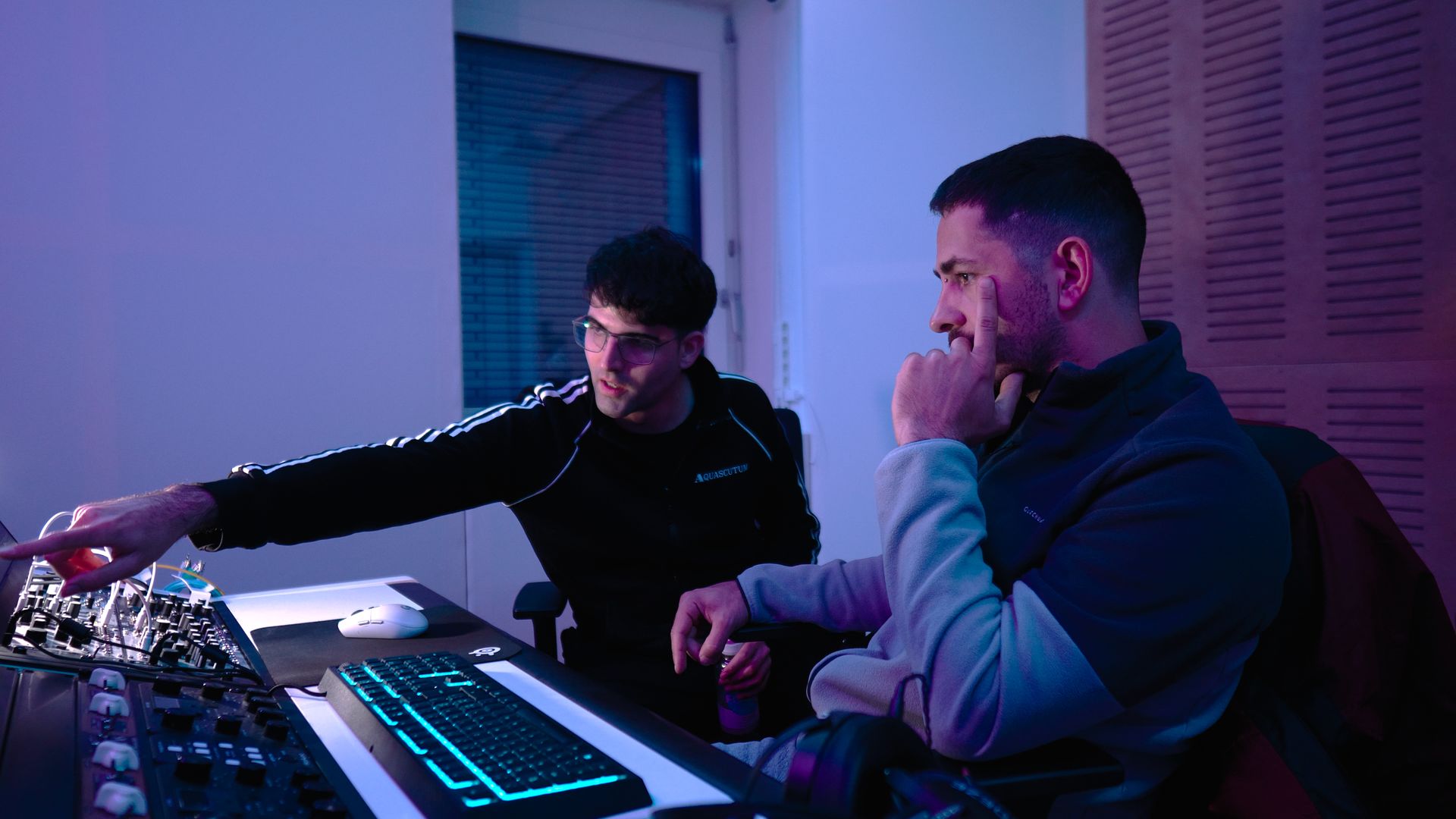 Zwei Personen mit Headsets arbeiten an einem Gaming-Setup und konzentrieren sich auf Bildschirme in violett-blauer Beleuchtung.