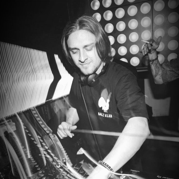 DJ in a club, long hair, black shirt, headphones, mixing music at a console.