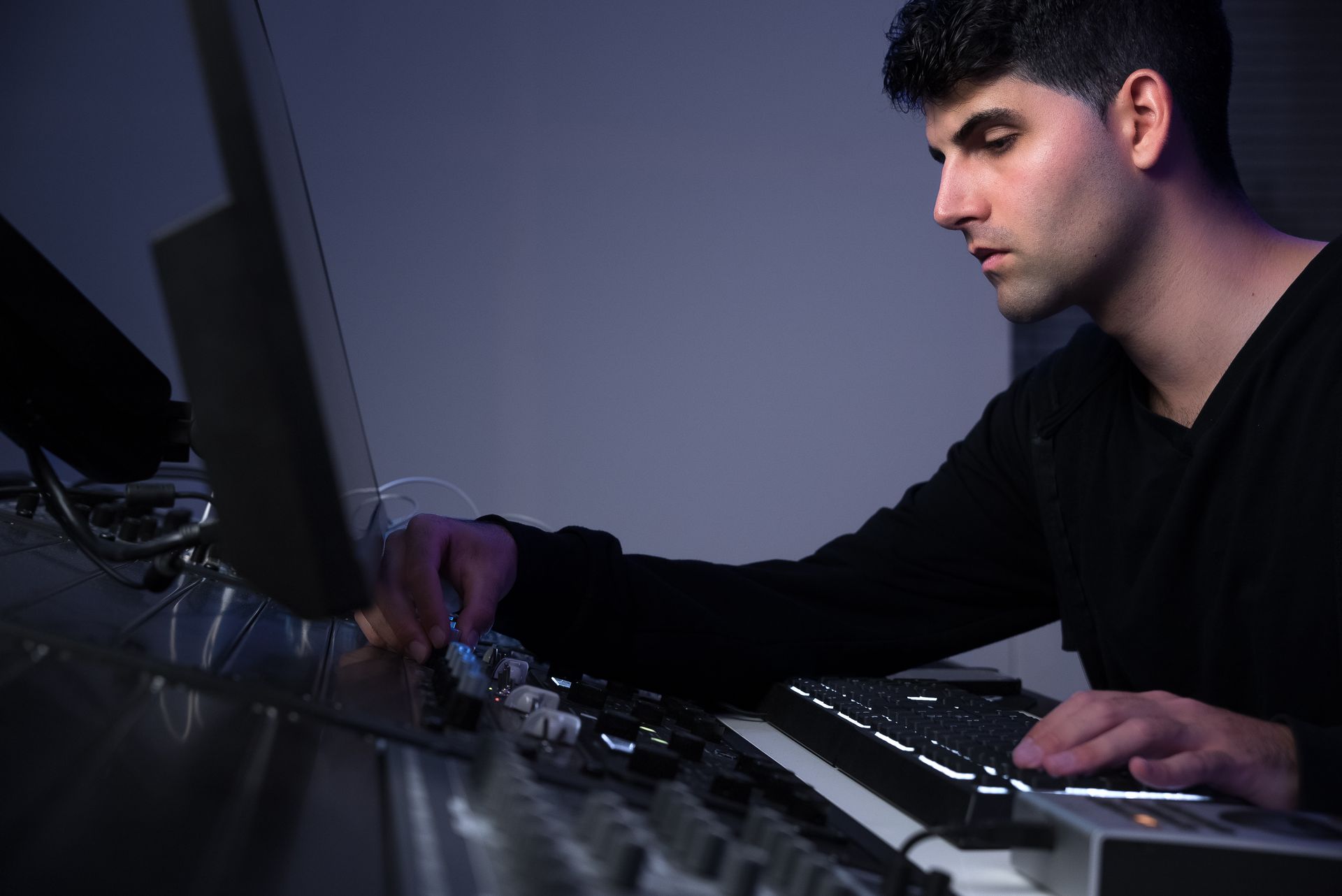 Man in black sweater adjusts knobs on a keyboard, working at a computer in a dimly lit room.